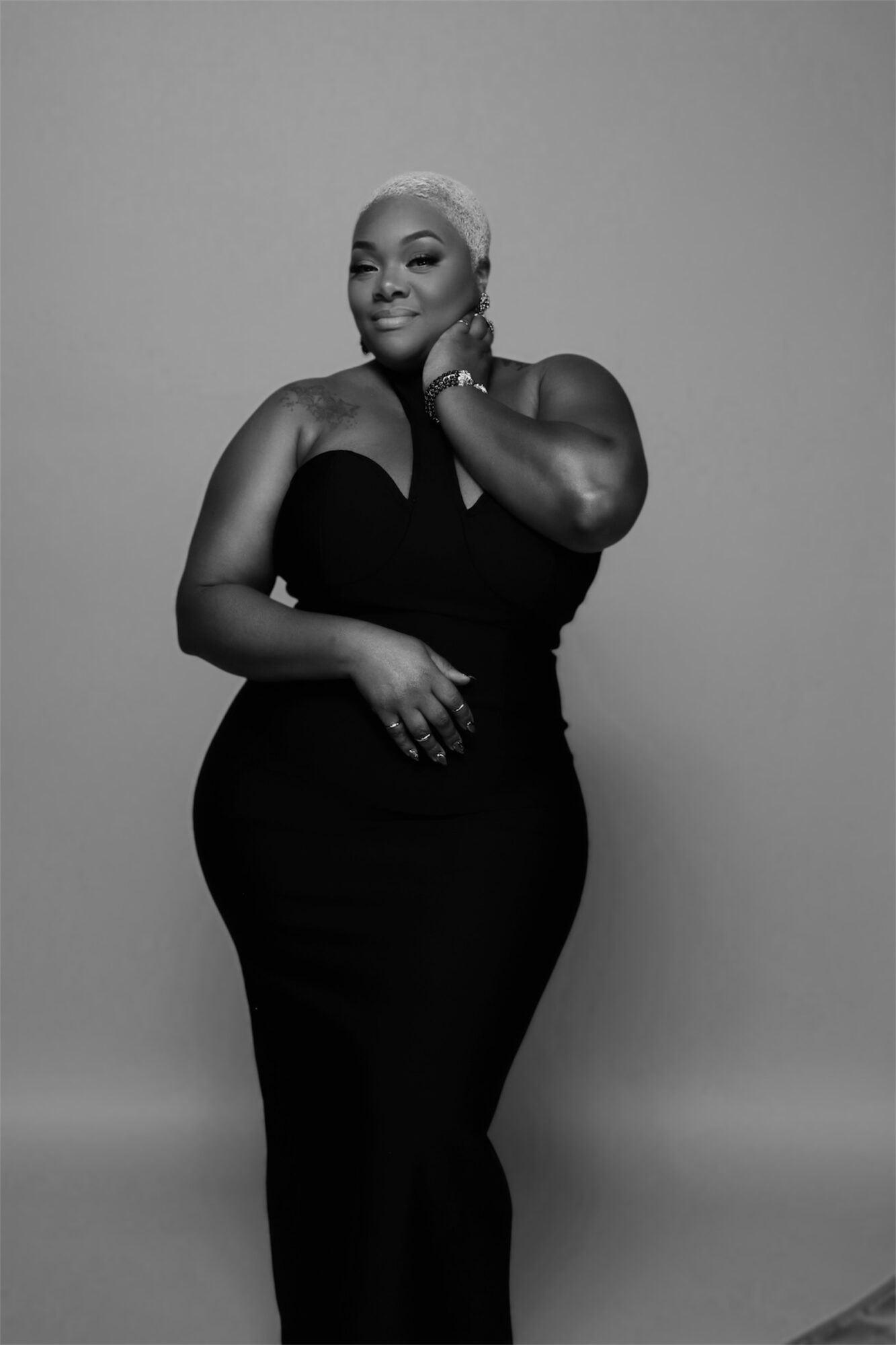 Woman with short hair in a strapless dress, smiling, posing with hand near face, against plain background.