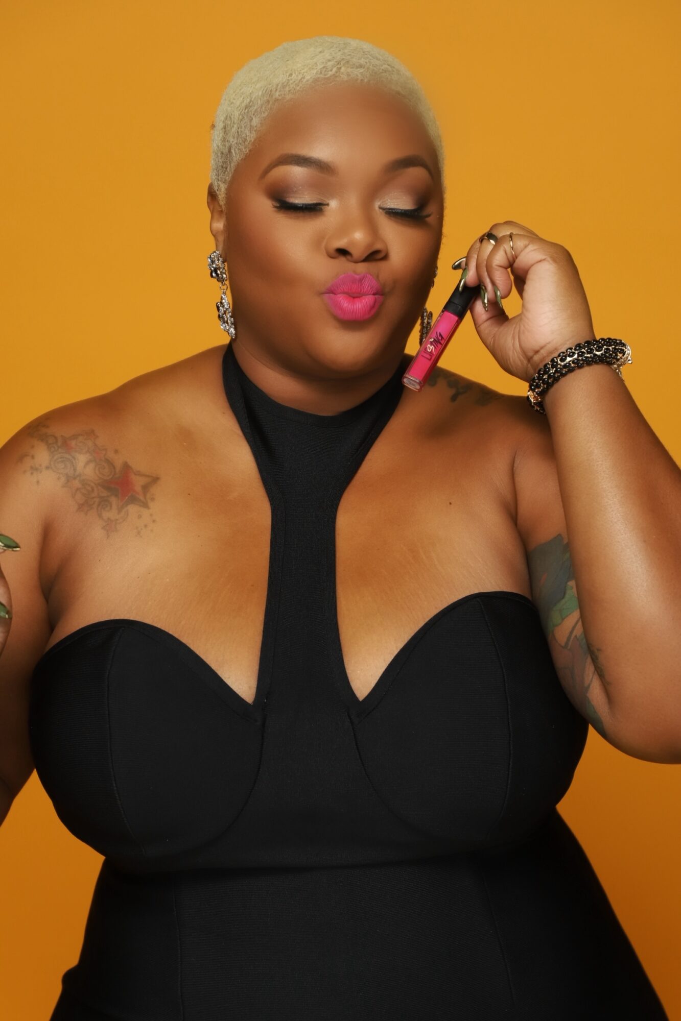 Woman with short blonde hair holding lipstick near face, wearing black dress and jewelry, against orange background.