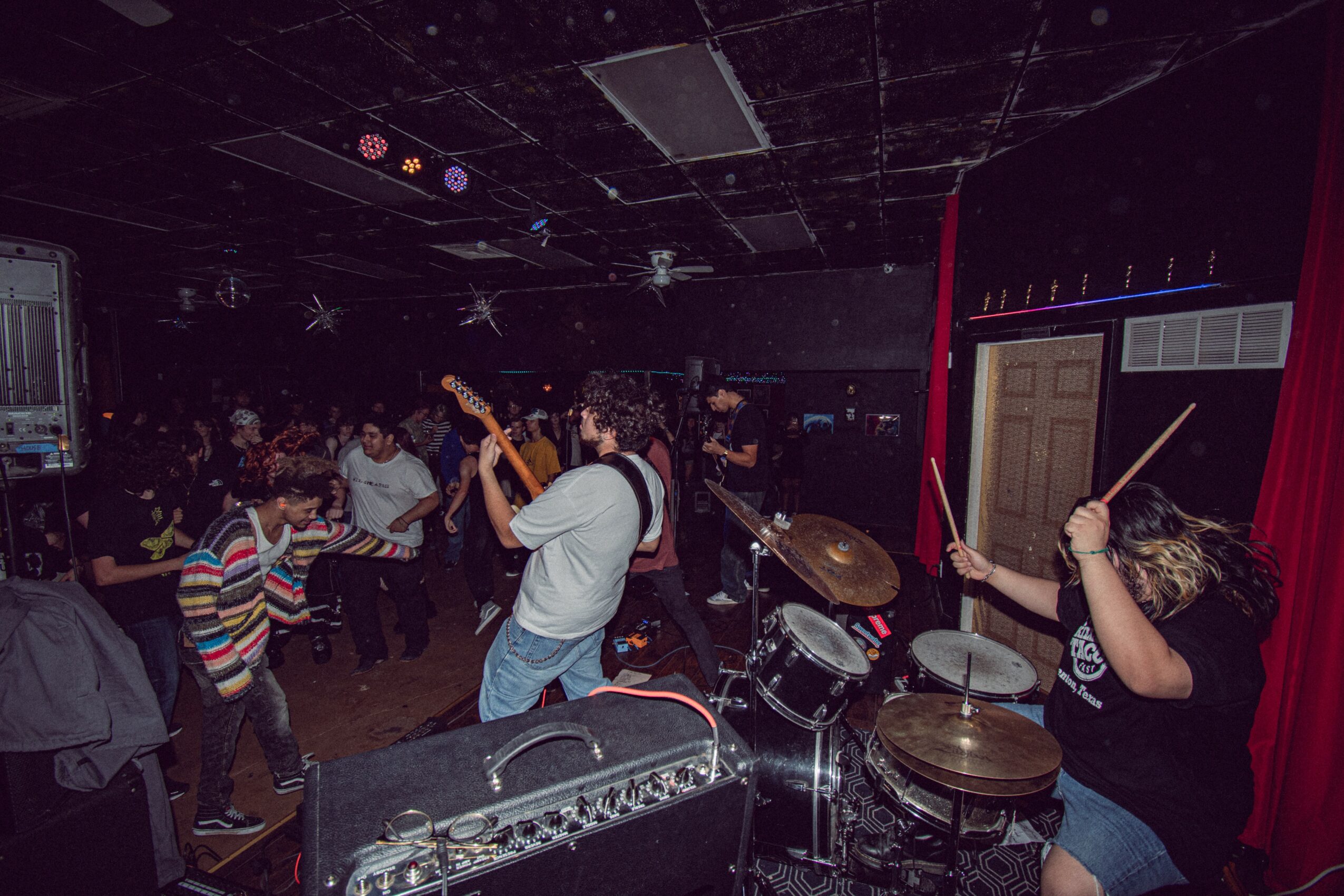 People dancing and playing drums in a dark indoor space with stage curtains and equipment.