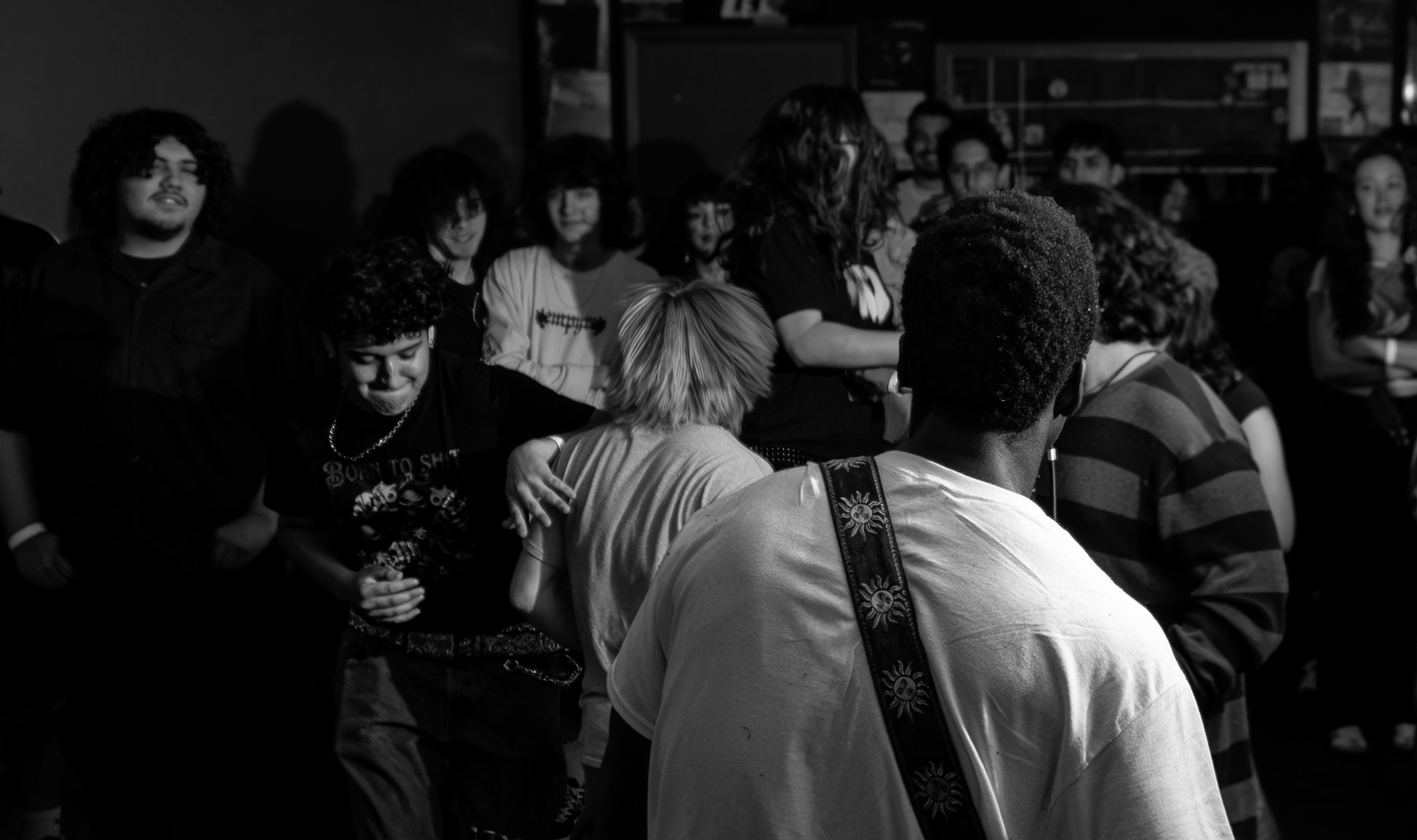Group of people gathered indoors, some facing forward, others turned, with shelves and objects in background.
