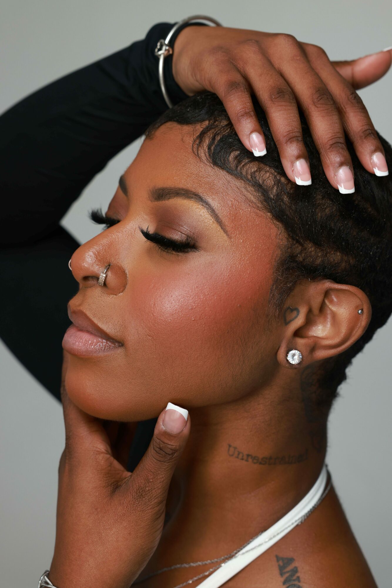 Close-up of a woman with eyes closed, touching her forehead and neck, with makeup and jewelry visible.