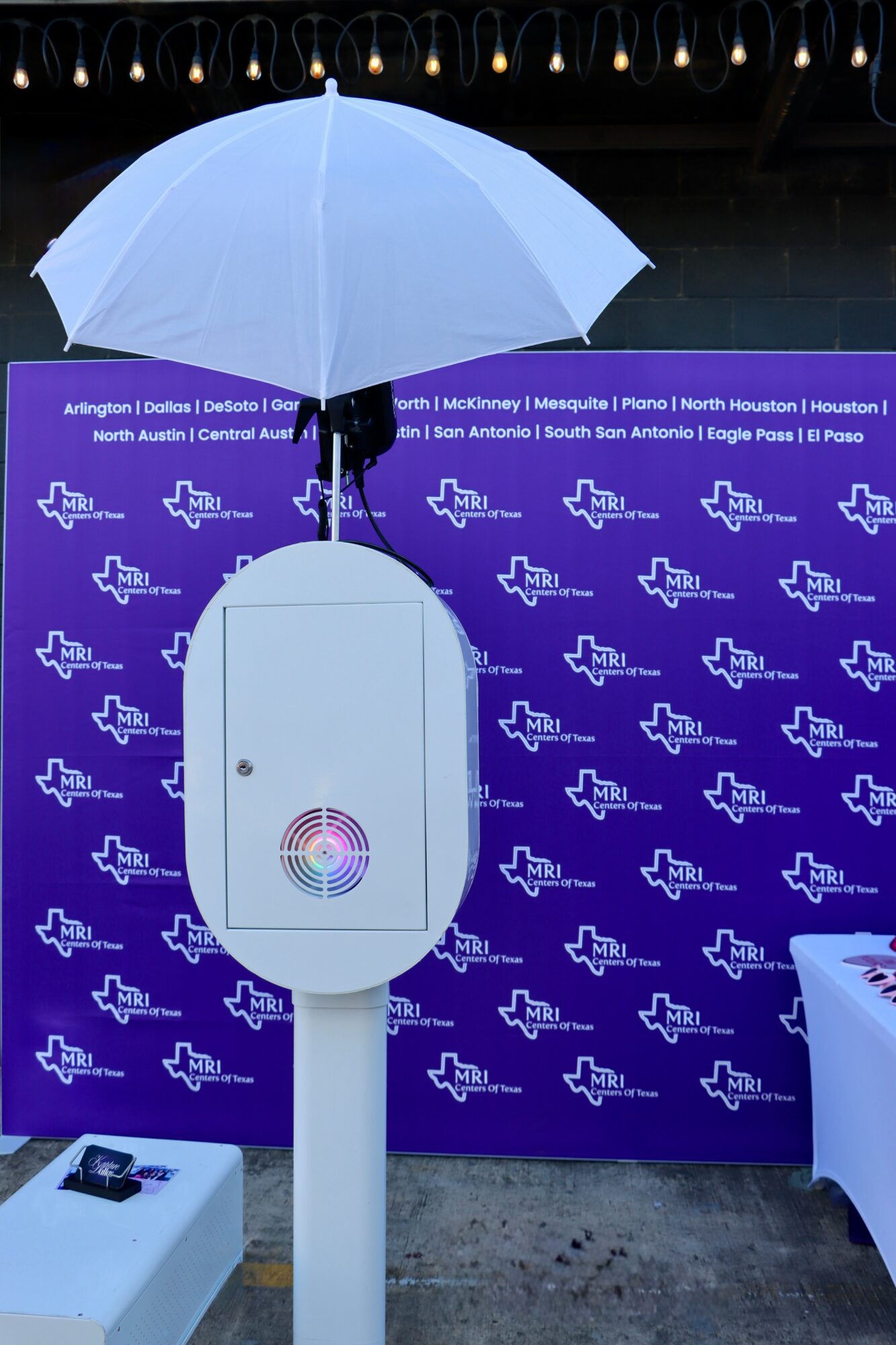 White umbrella with a purple banner behind it, displaying text and logos, set against a dark background with string lights.