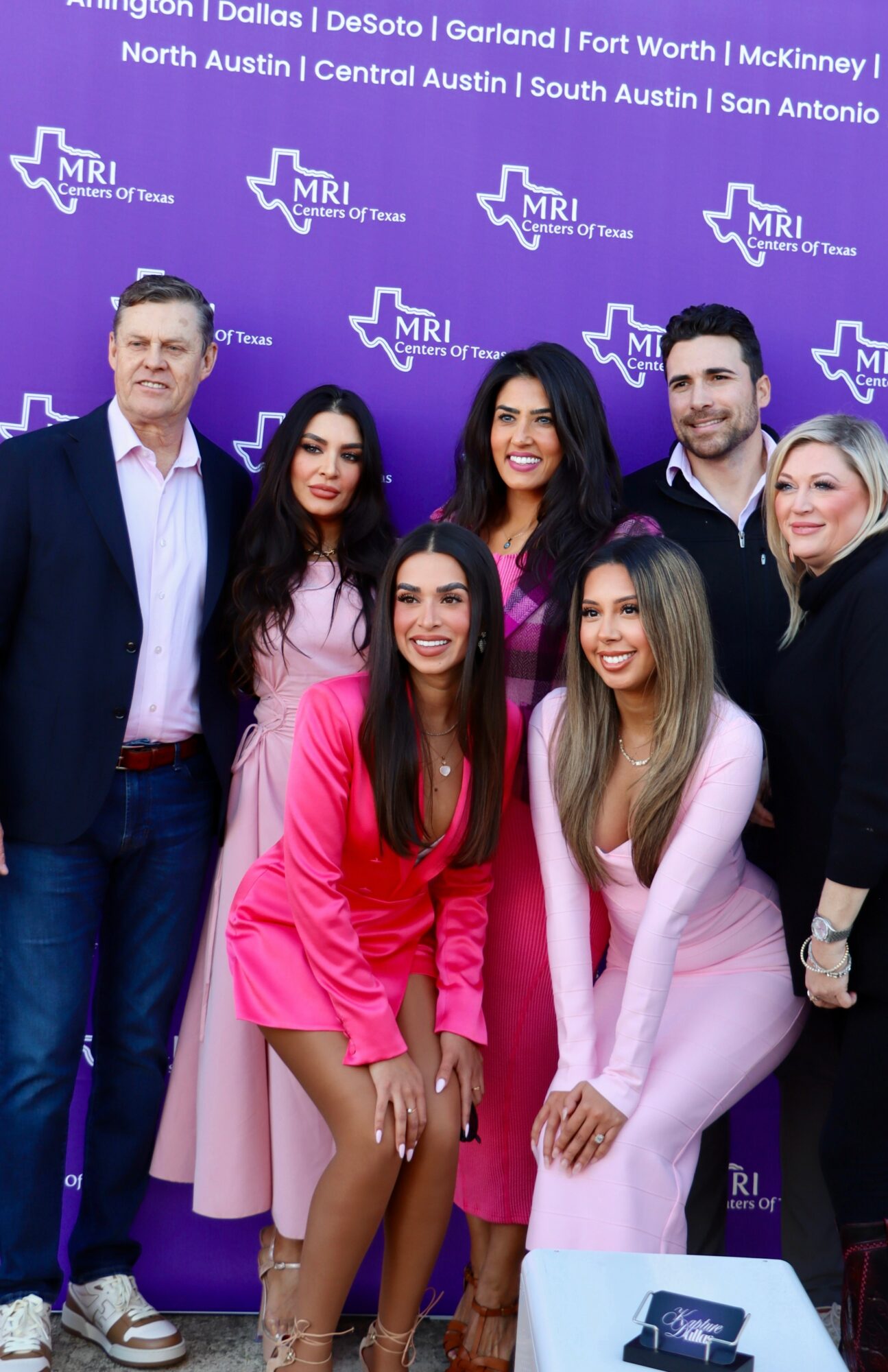 Group of six people standing in front of a purple backdrop with Texas map and city names, smiling for photo.