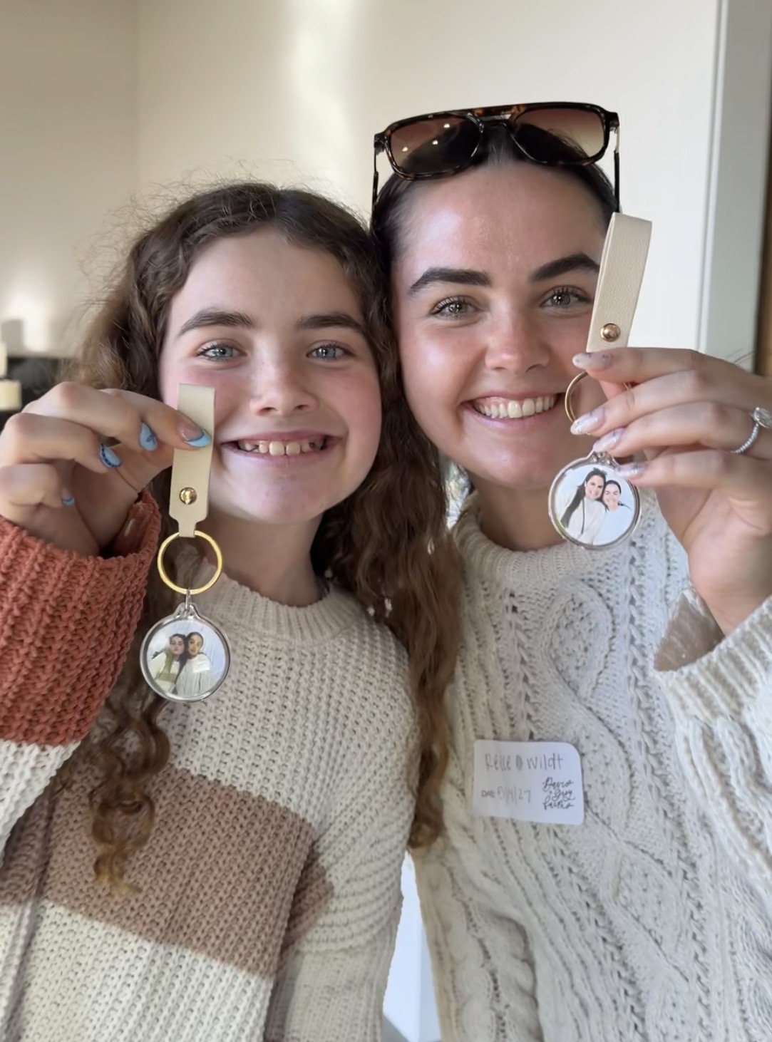 Two smiling women holding keychains with photos, standing indoors, wearing sweaters, one with sunglasses on head.