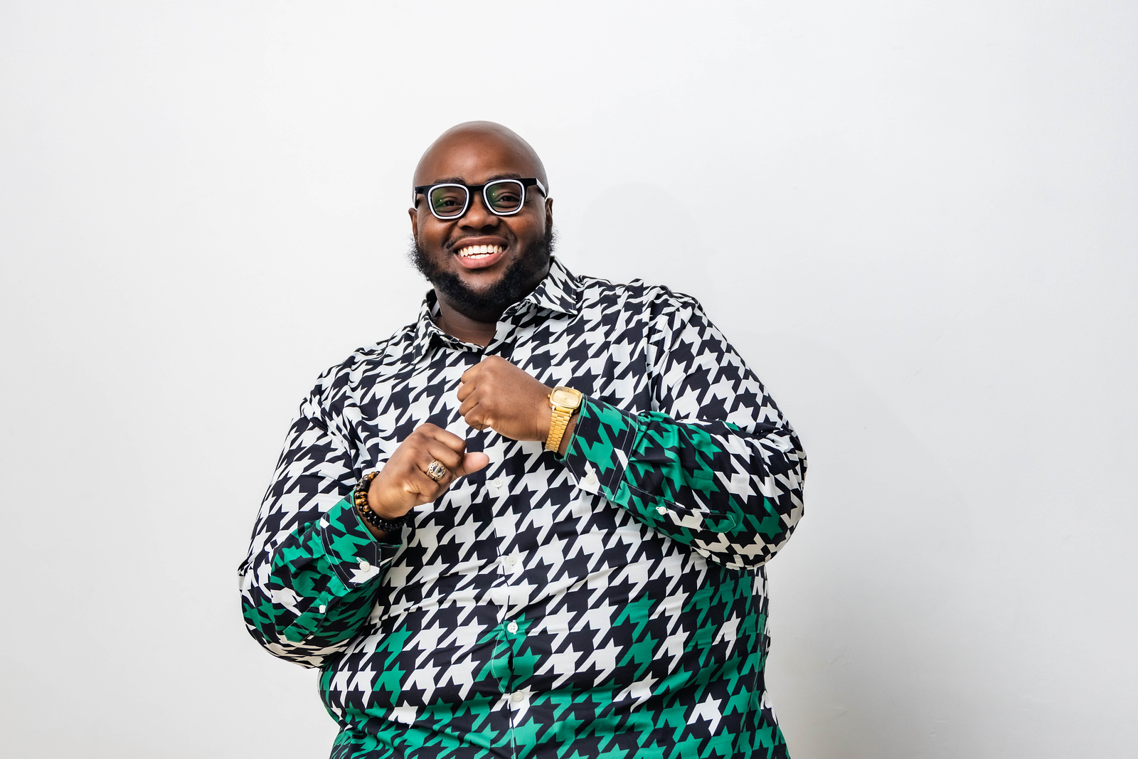 Smiling man with glasses wearing a black and white houndstooth shirt and green patterned sleeves, standing against a white background.