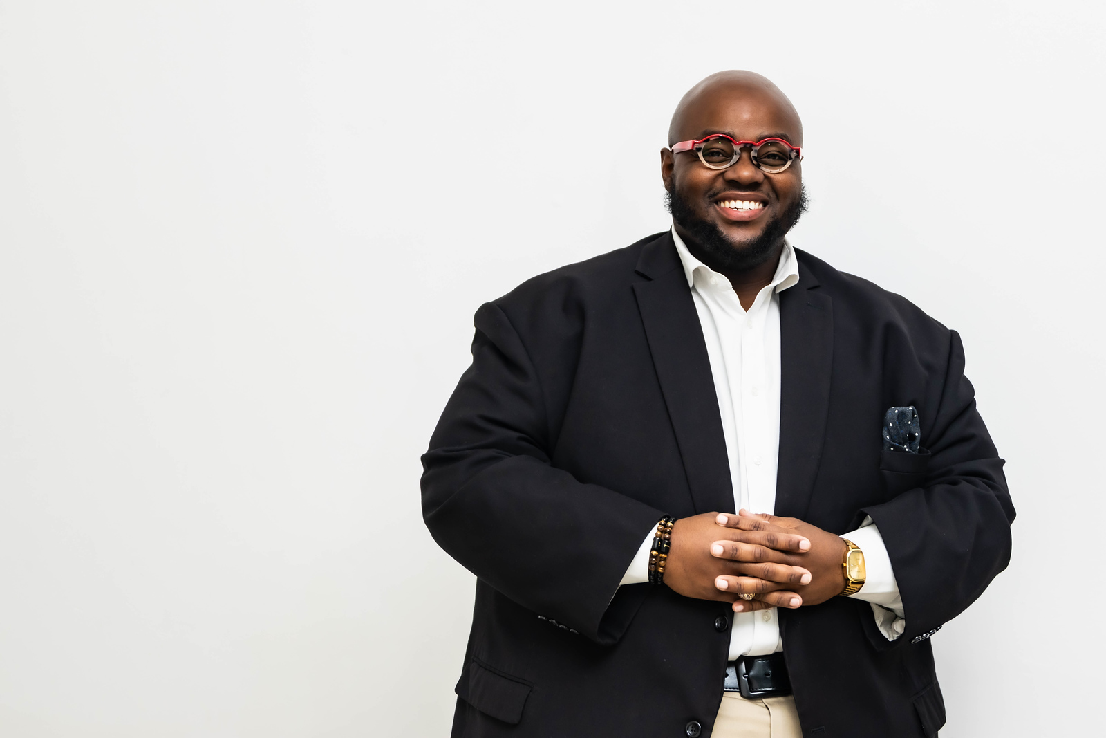 Man with glasses and beard in a suit, smiling, standing with hands clasped, against a plain white background.