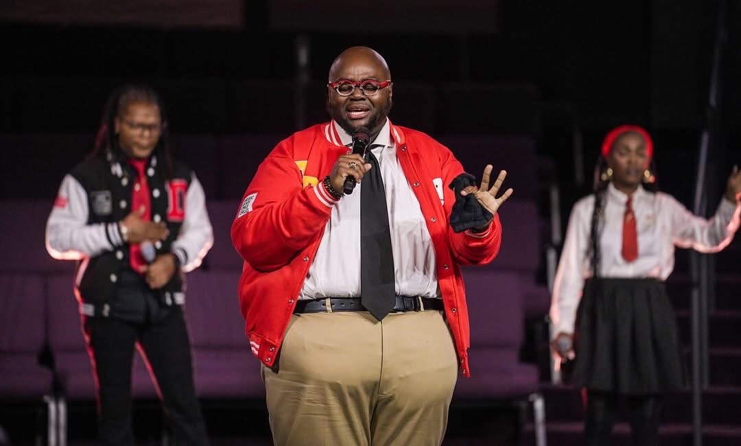 Man speaking into microphone on stage with two girls in school uniforms behind him.