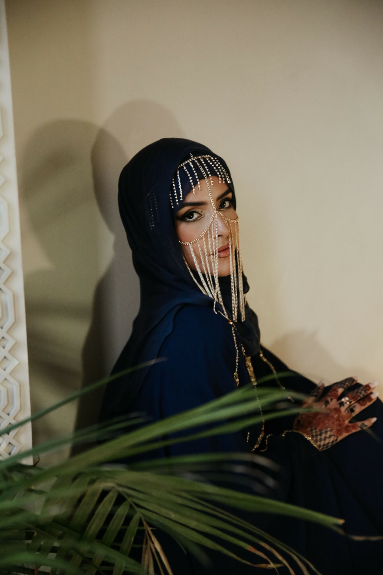 Woman wearing a dark headscarf with decorative beads, sitting indoors with a neutral wall background.