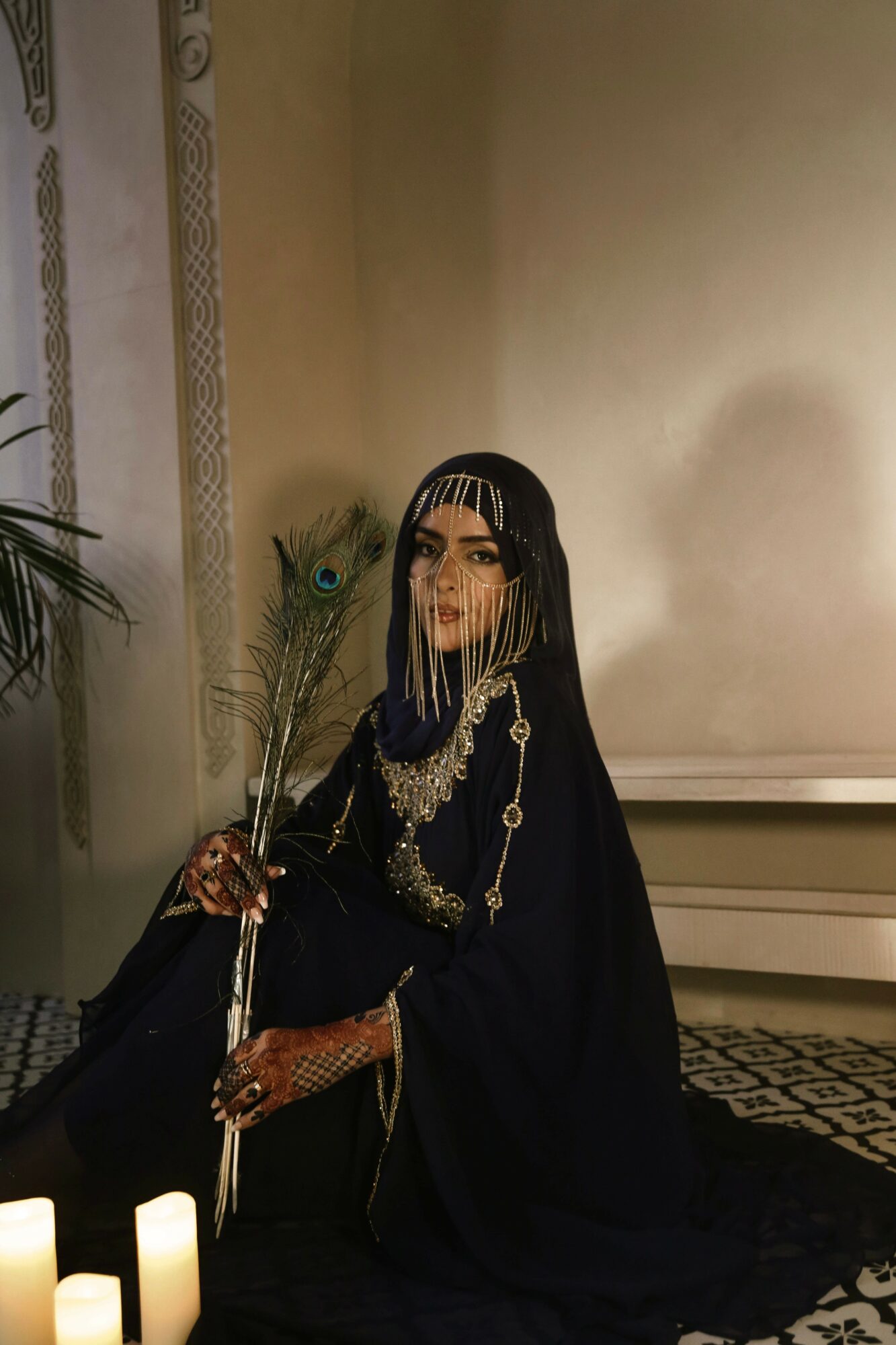 Woman in dark clothing with jewelry, sitting on a patterned floor, holding a peacock feather, in a room with beige walls.