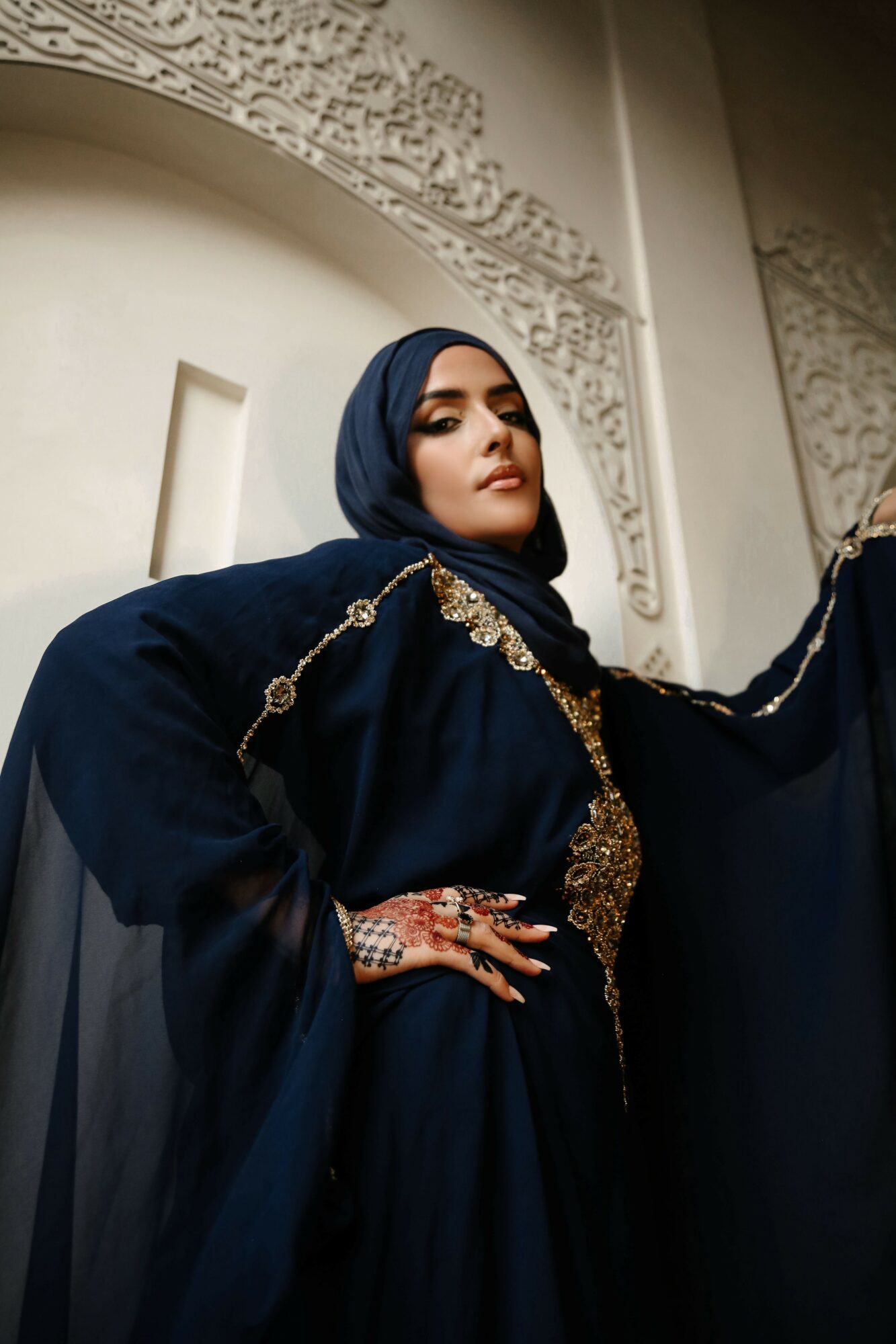 Woman in dark blue traditional attire with gold embellishments, wearing a headscarf, standing indoors with ornate decor.