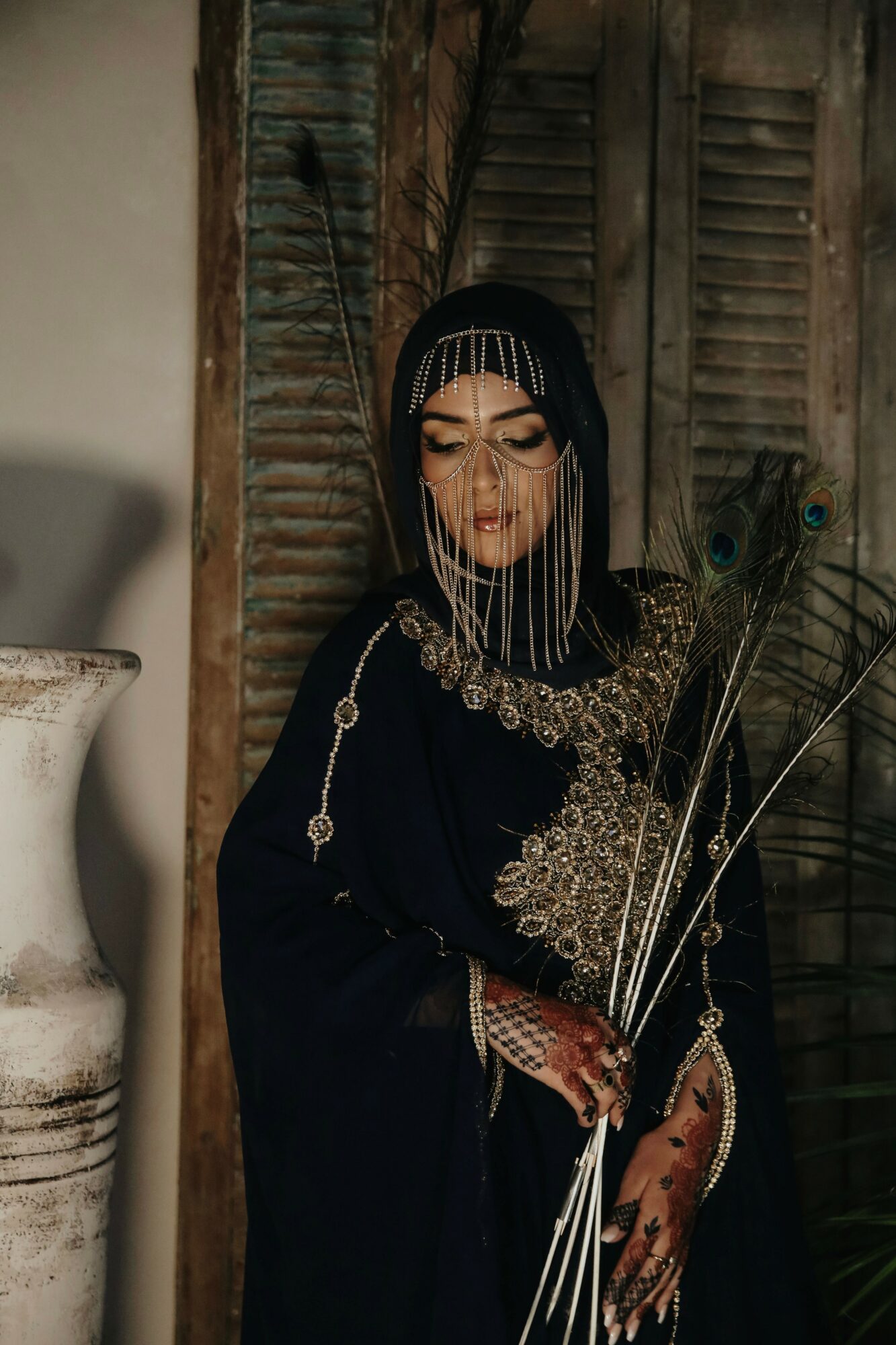 Woman in traditional attire with jewelry and henna, holding plants, standing in front of wooden shutters.