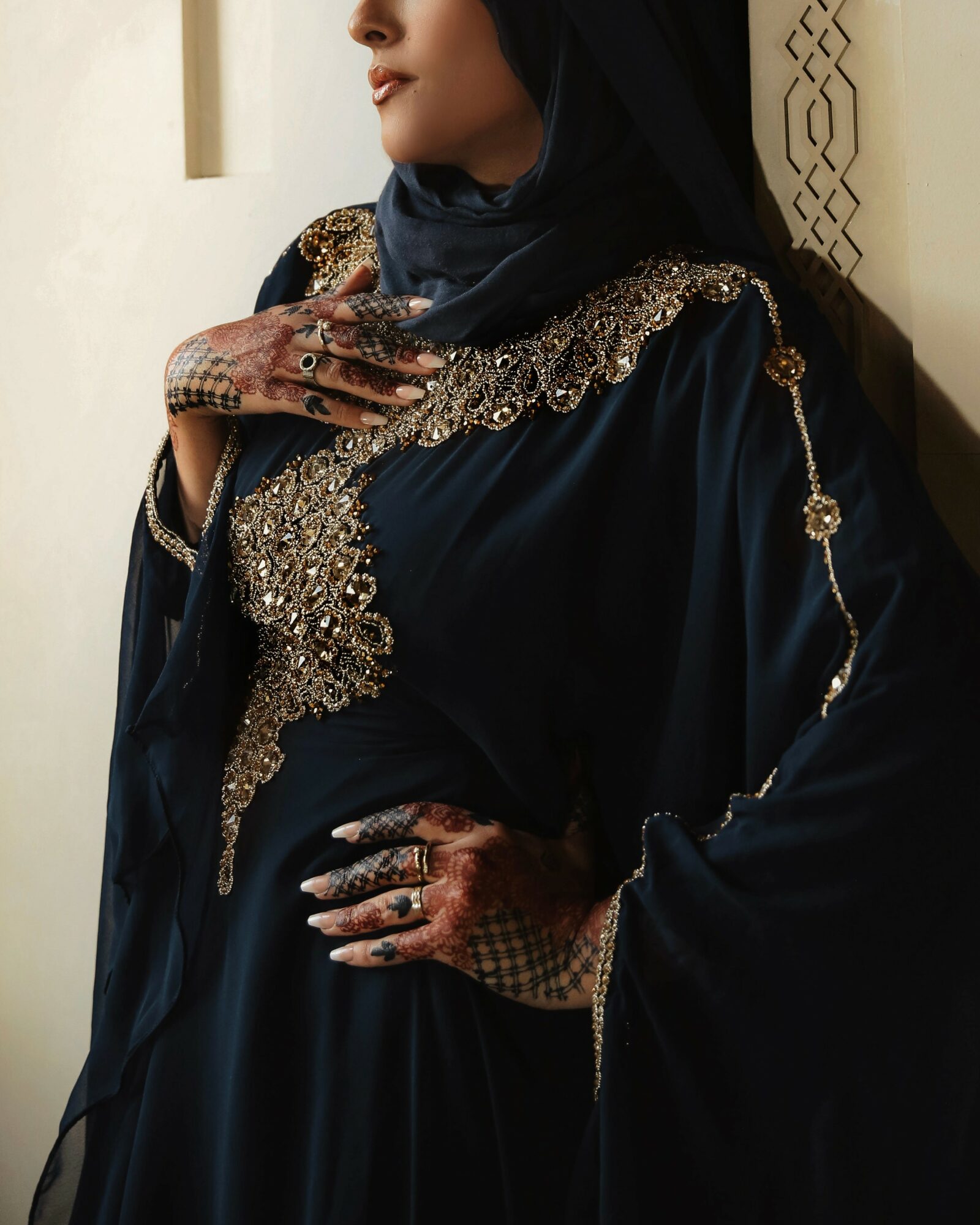 Woman in dark clothing with intricate gold embroidery, wearing rings and henna, standing against a light wall.