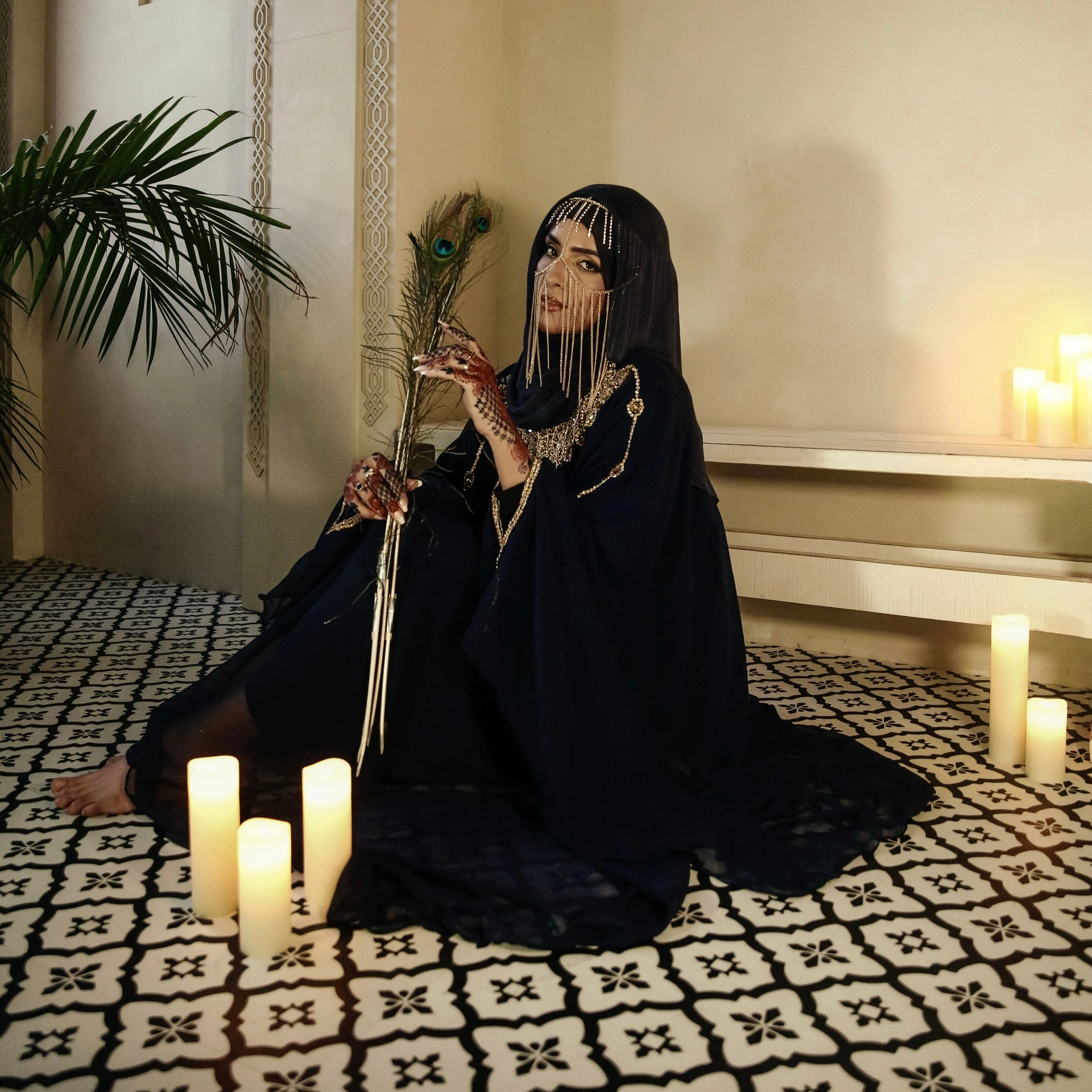 Woman in black dress sitting on patterned floor with candles, holding a peacock feather, in a decorated room.