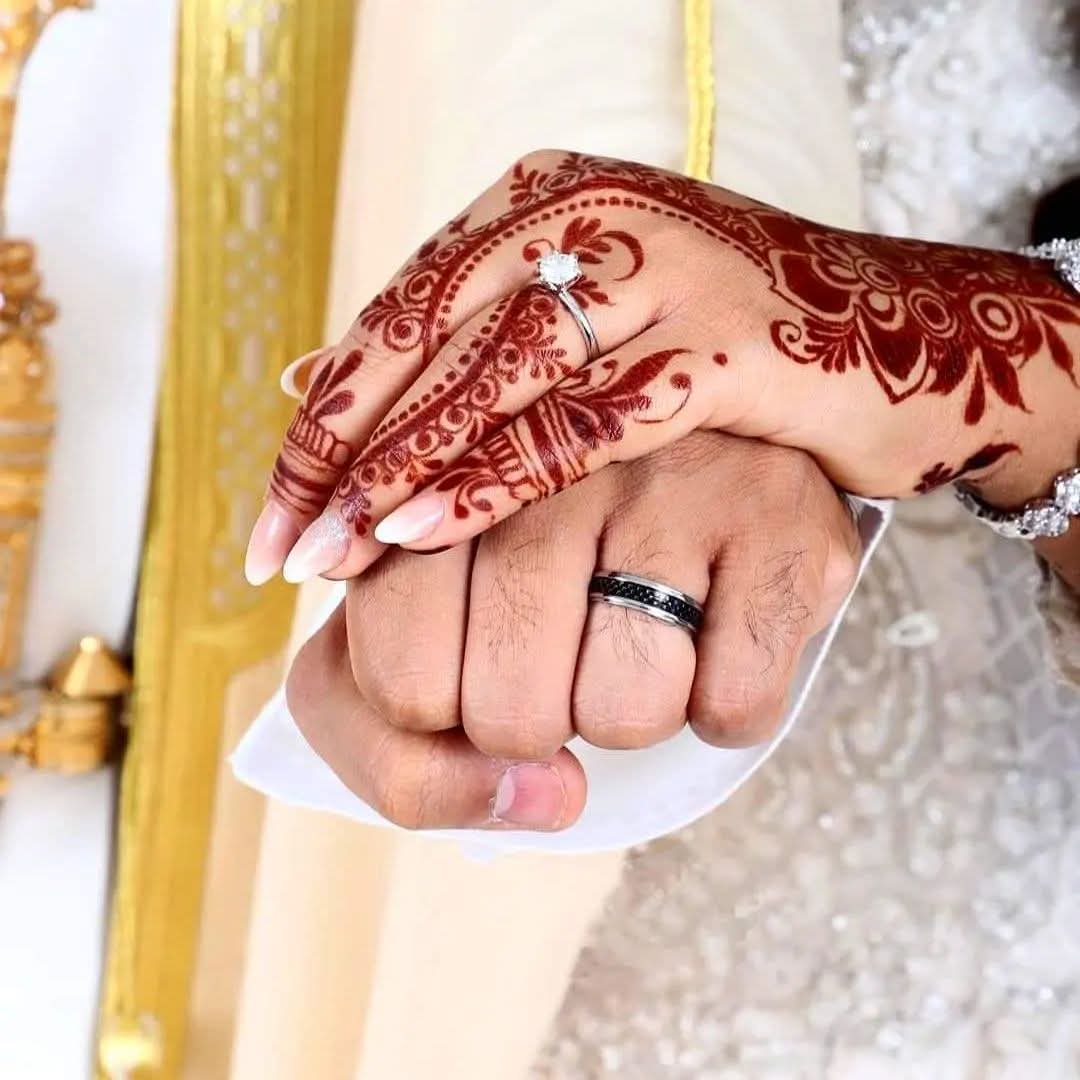 Two hands with henna designs holding each other, one with a wedding ring, background with yellow and white fabric.