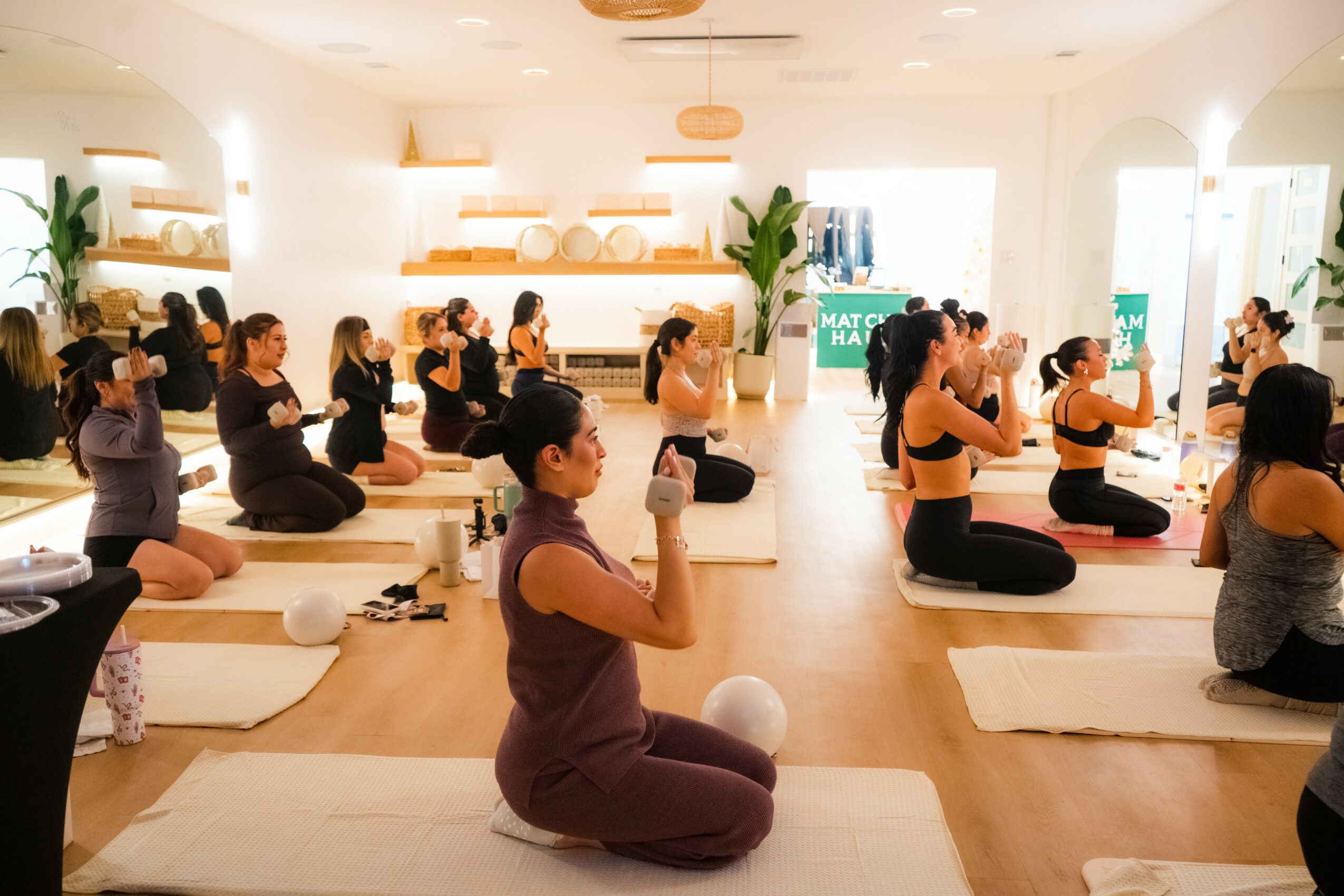 Group of people practicing yoga in a bright studio, sitting on mats with hands in prayer position, facing instructor.