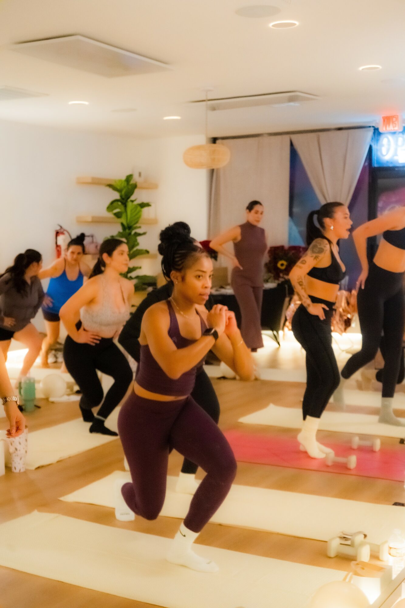 Group of women participating in a fitness class indoors, some in yoga poses, with mats on the floor and a plant in the background.