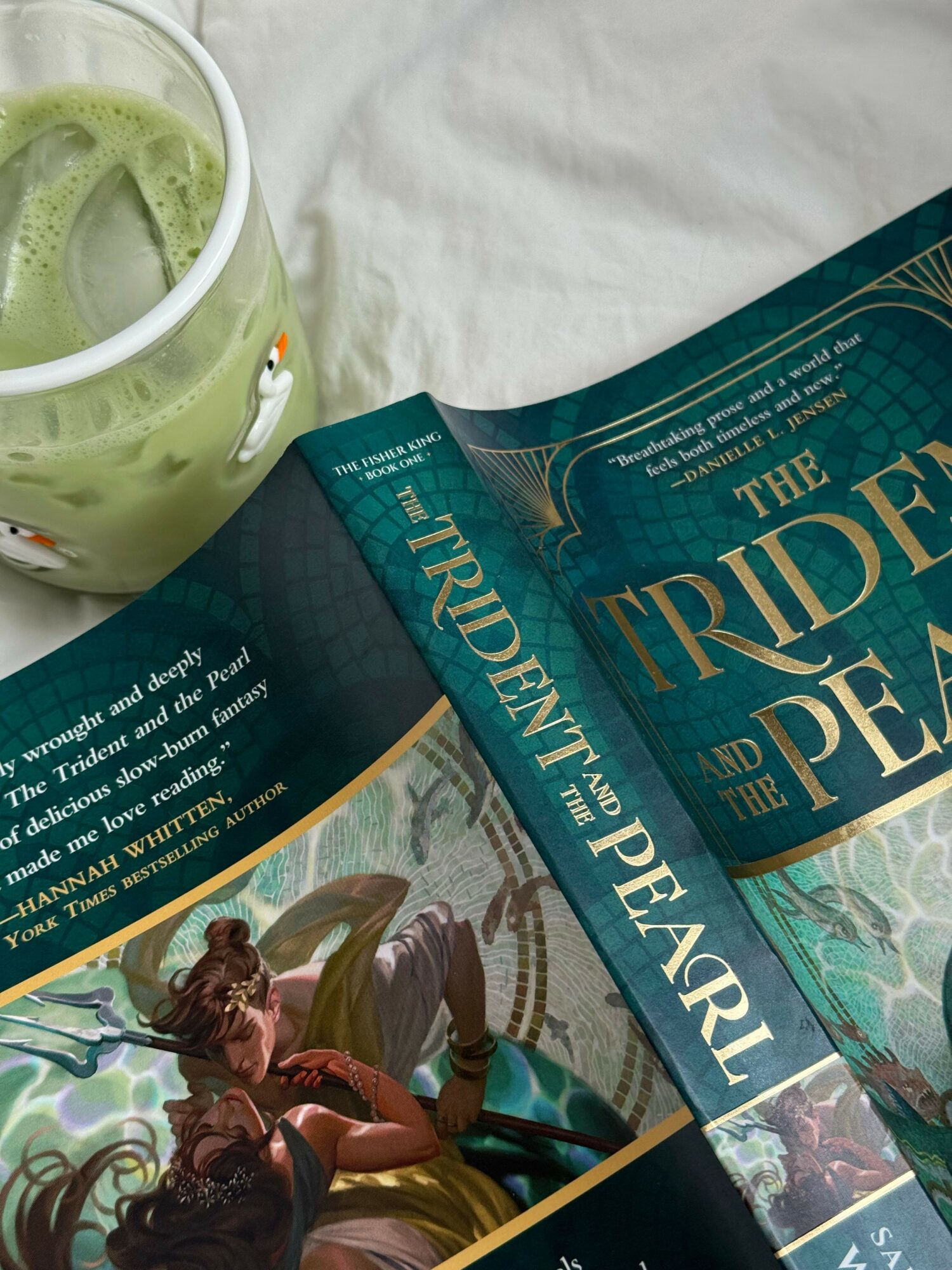 Green smoothie in a mug with a cartoon character, open book titled 'The Triumph of the Pearl' on a white cloth surface.