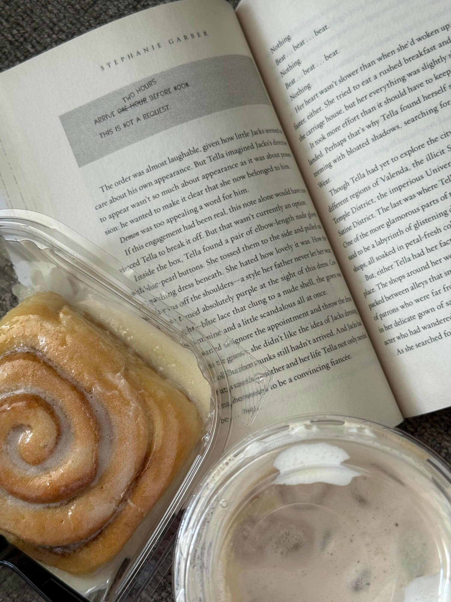 Open book with text, cinnamon roll in plastic container, and a cup of coffee or tea.
