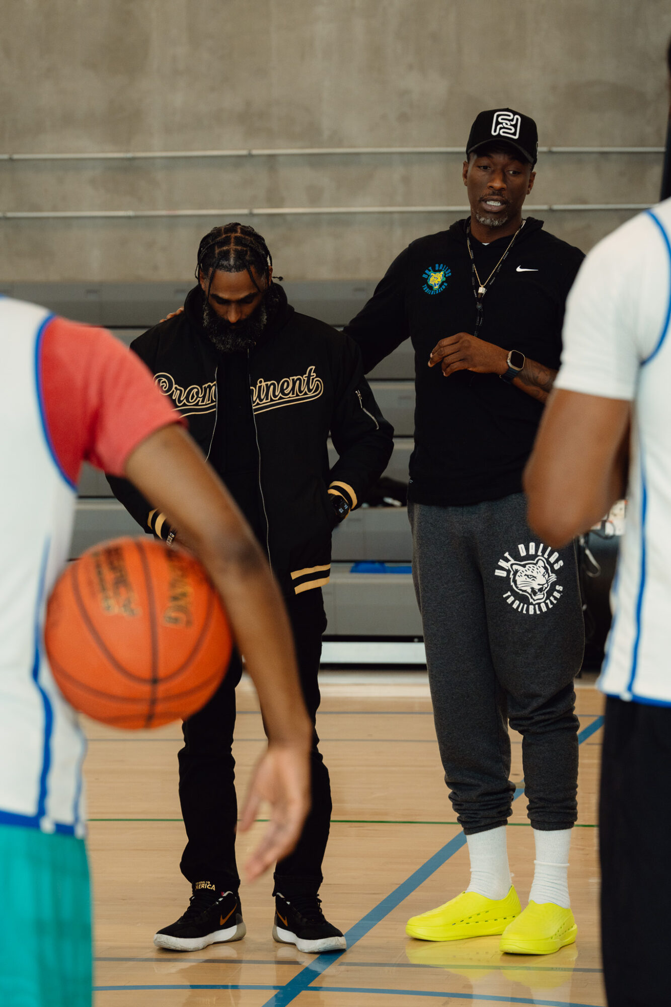 Two people standing on a basketball court, one wearing a black jacket and the other in a black hoodie, with a basketball nearby.