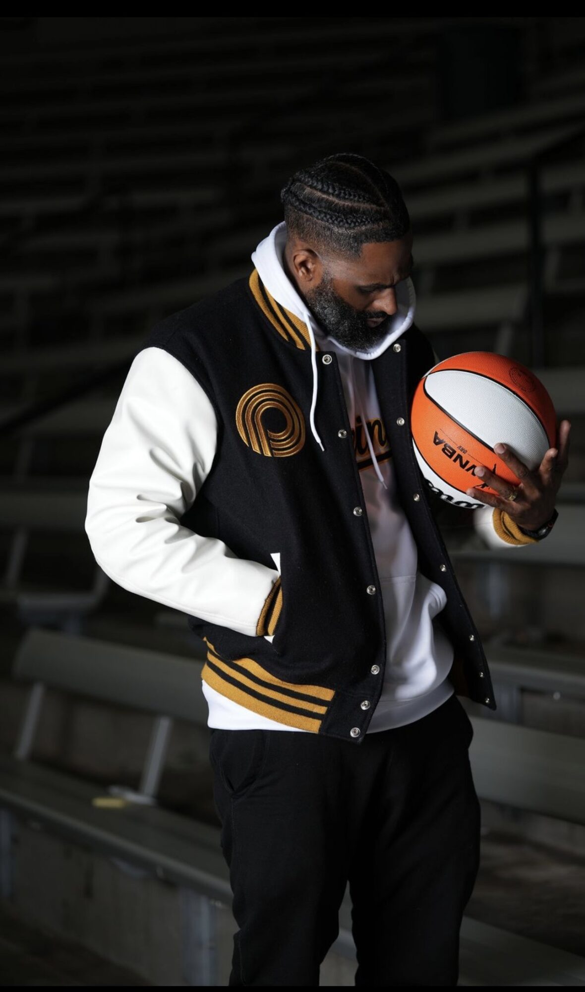 Man holding a basketball, standing in an empty stadium, wearing a varsity jacket and hoodie, looking down at the ball.