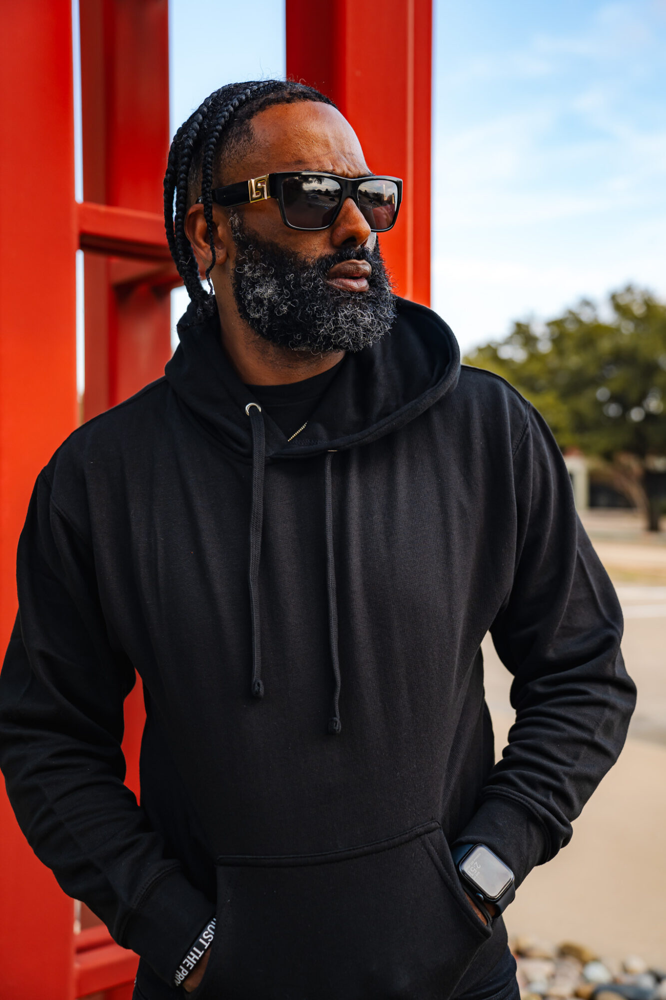 Man with sunglasses, beard, and braided hair wearing a black hoodie outdoors near a red structure.