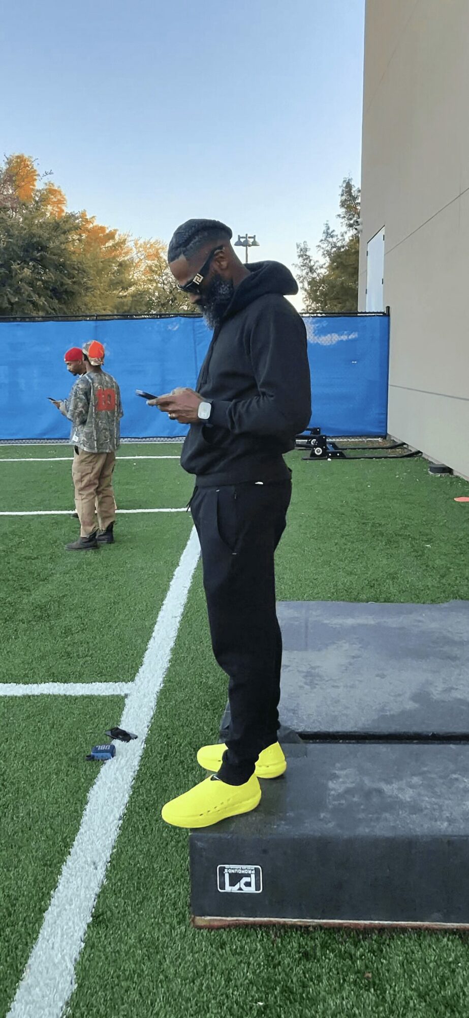 Young man in black hoodie and pants standing on a platform outdoors, looking at his phone, with a person in the background.