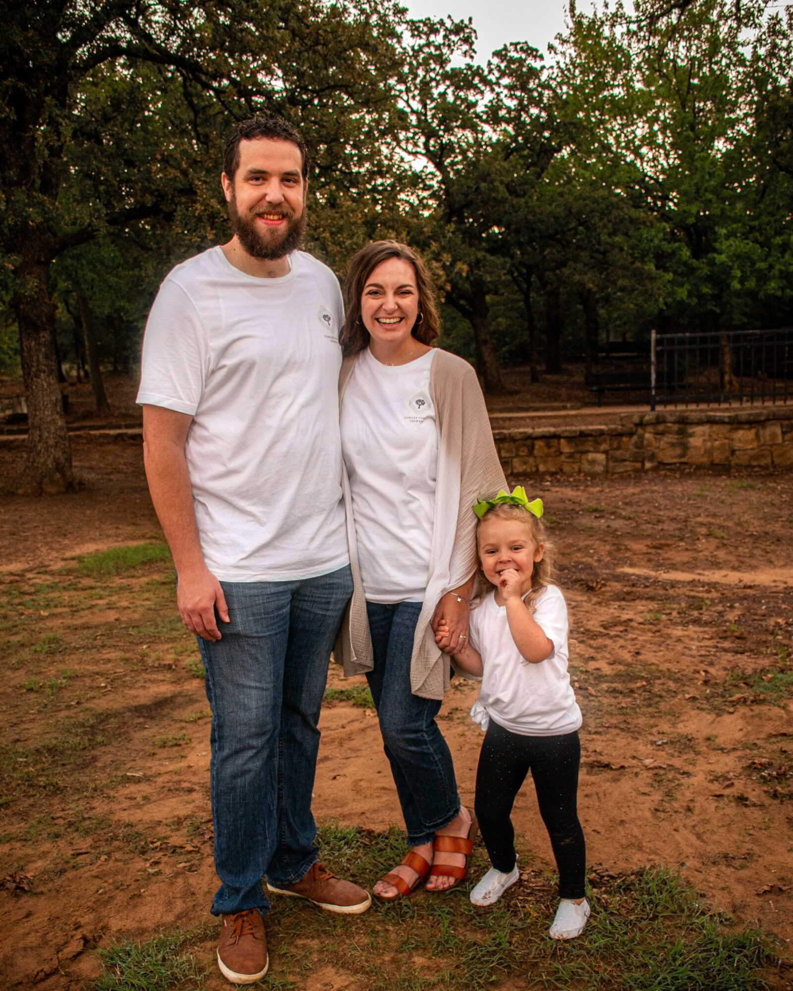 Three people, two adults and one child, standing outdoors on dirt ground with trees in background.