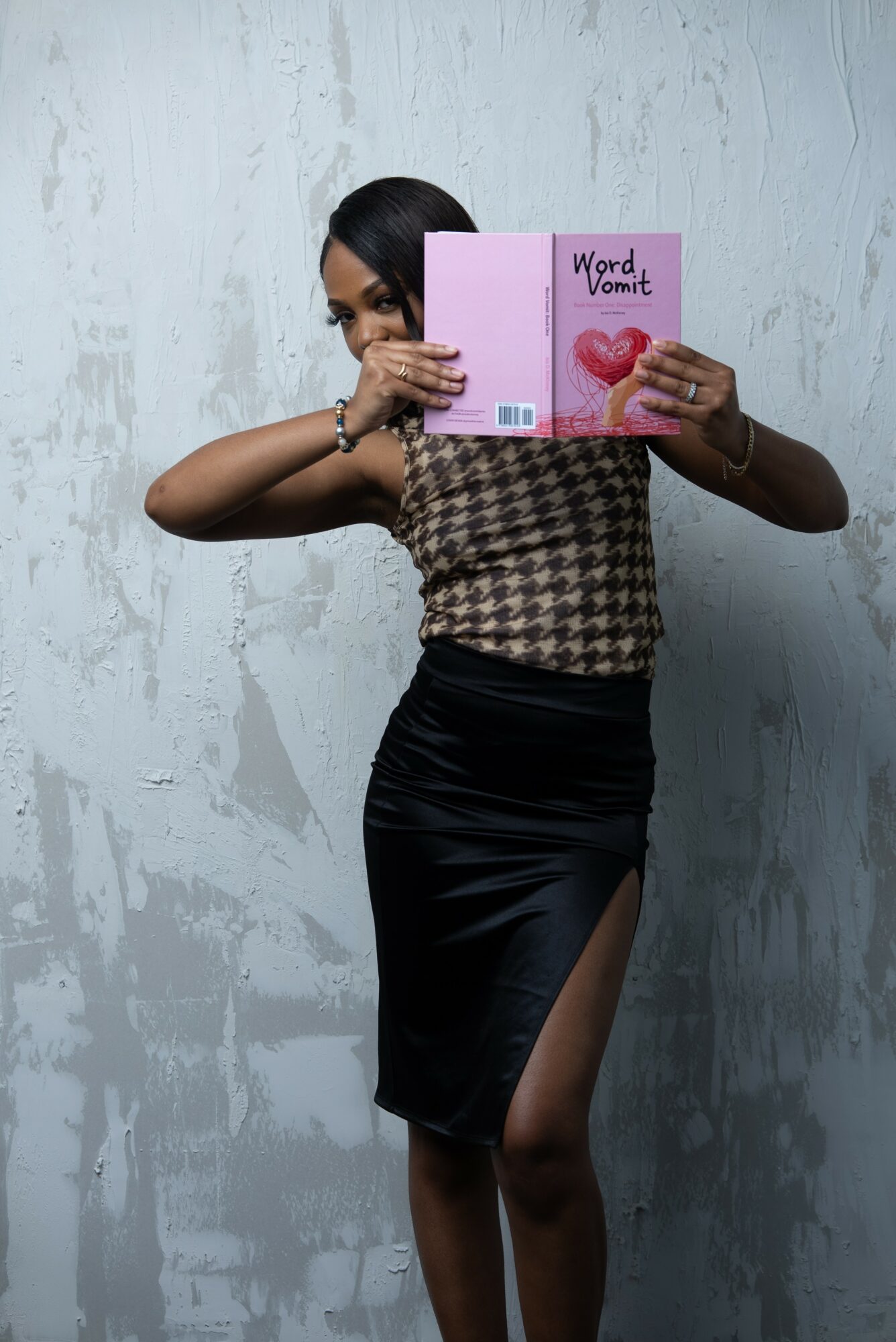Person holding a pink book titled 'Word Vomit' in front of face, standing against a textured white wall.