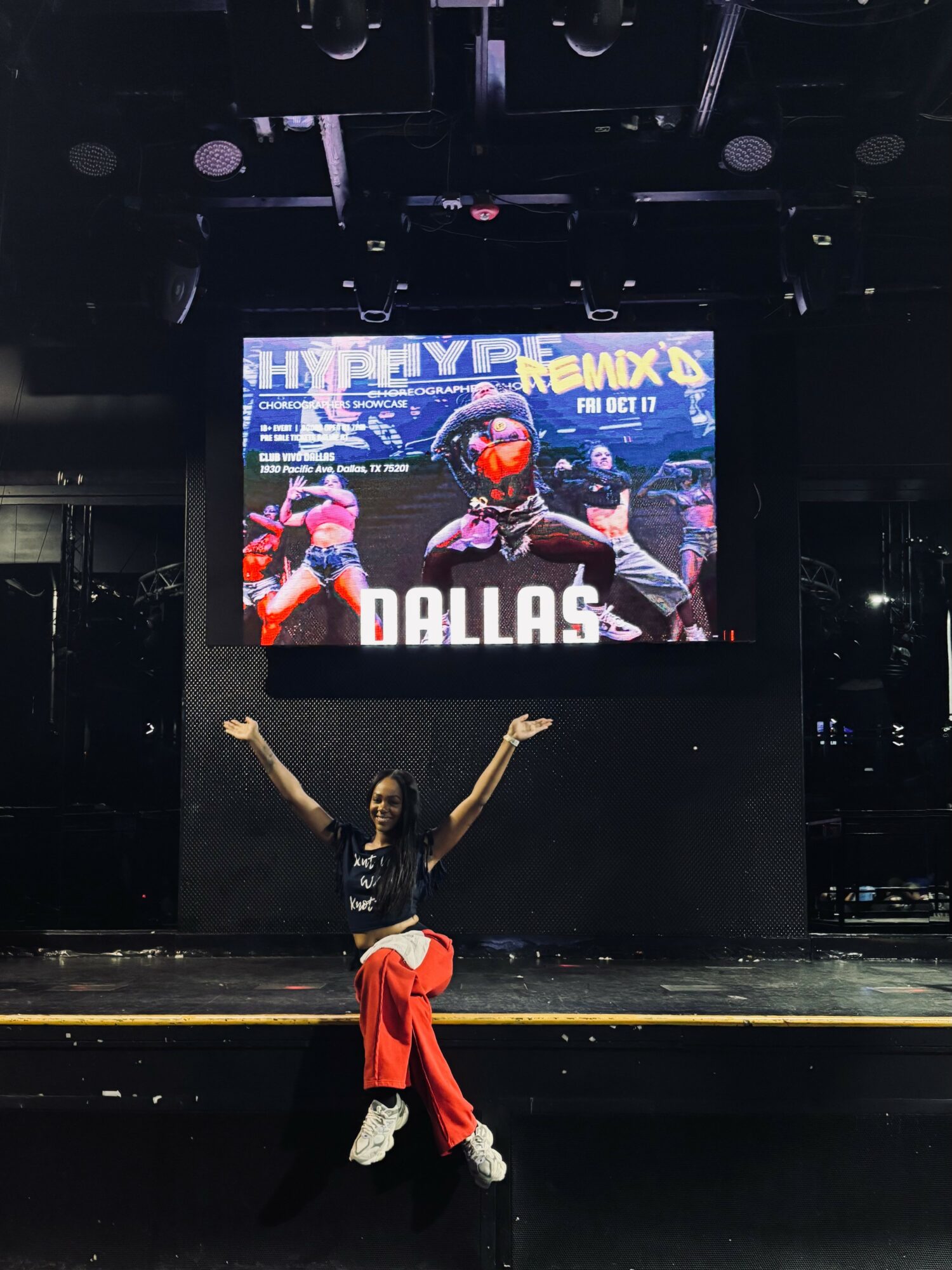 Person on stage with arms raised, wearing red pants and black top, in front of large screen displaying a racing scene.