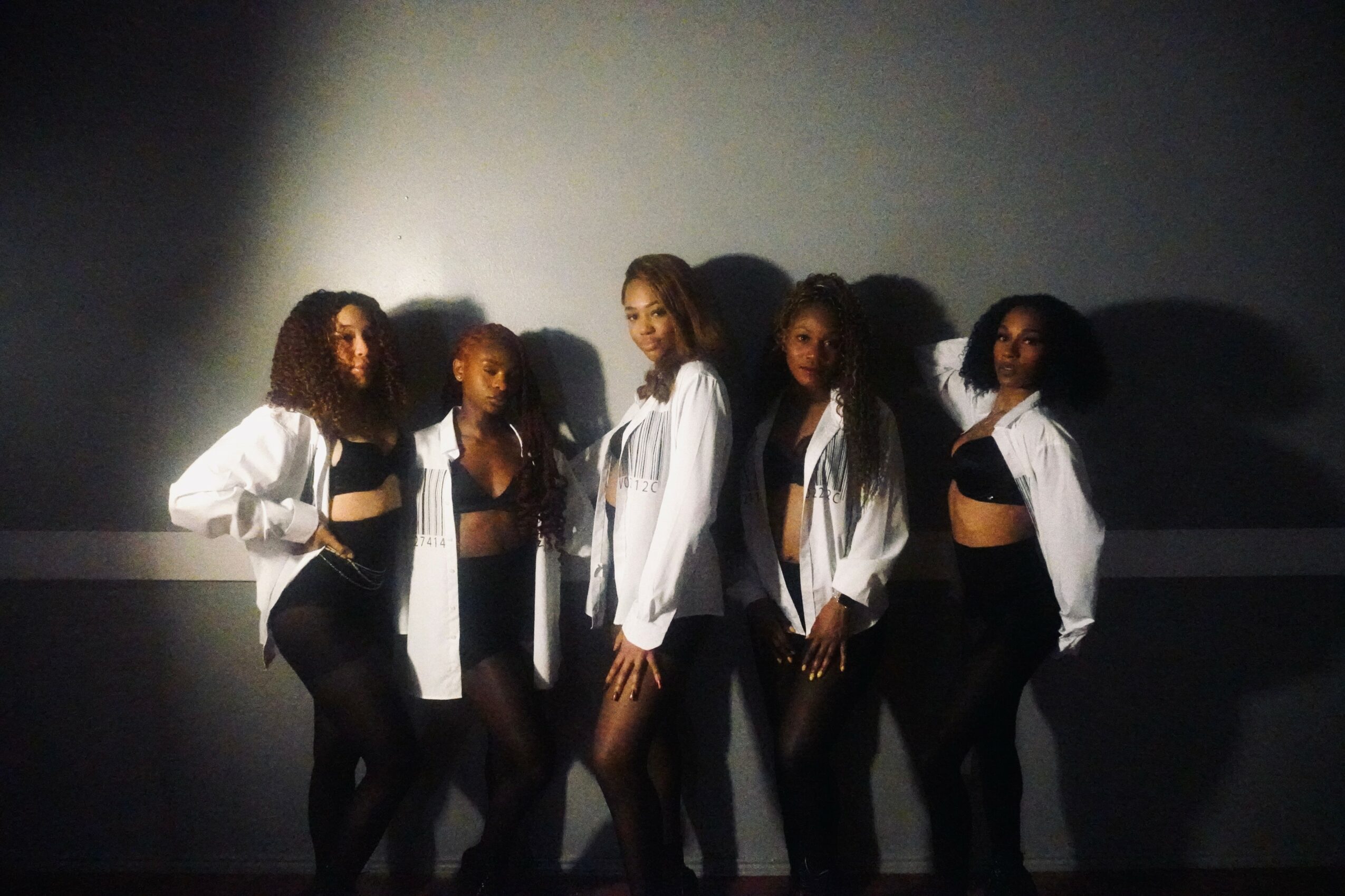 Five women standing against a plain wall, dressed in black and white outfits, with shadows cast behind them.