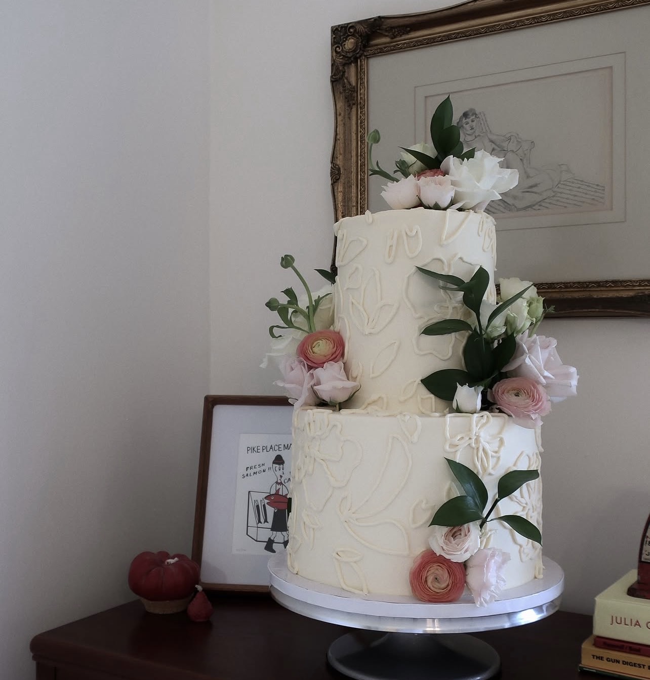 Two-tiered white cake decorated with pink and white flowers and green leaves, on a cake stand.