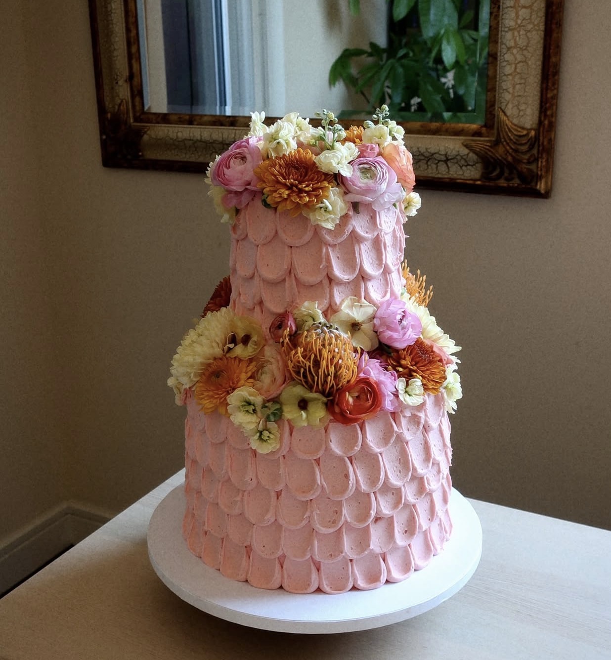 Two-tier pink cake decorated with flowers, on a white plate, in a room with a mirror and window.
