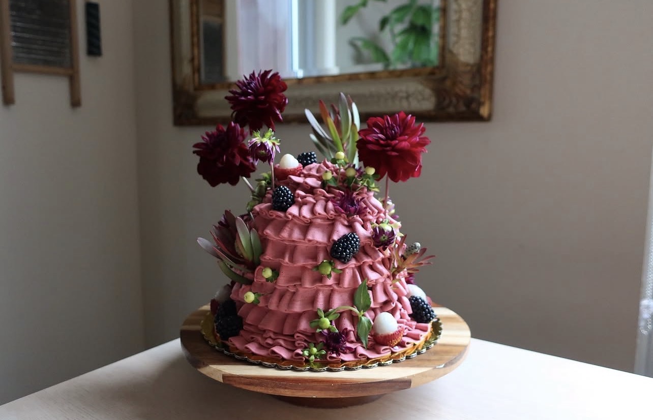Pink flower-adorned cake with berries and greenery on a wooden plate, in a room with a mirror and window.