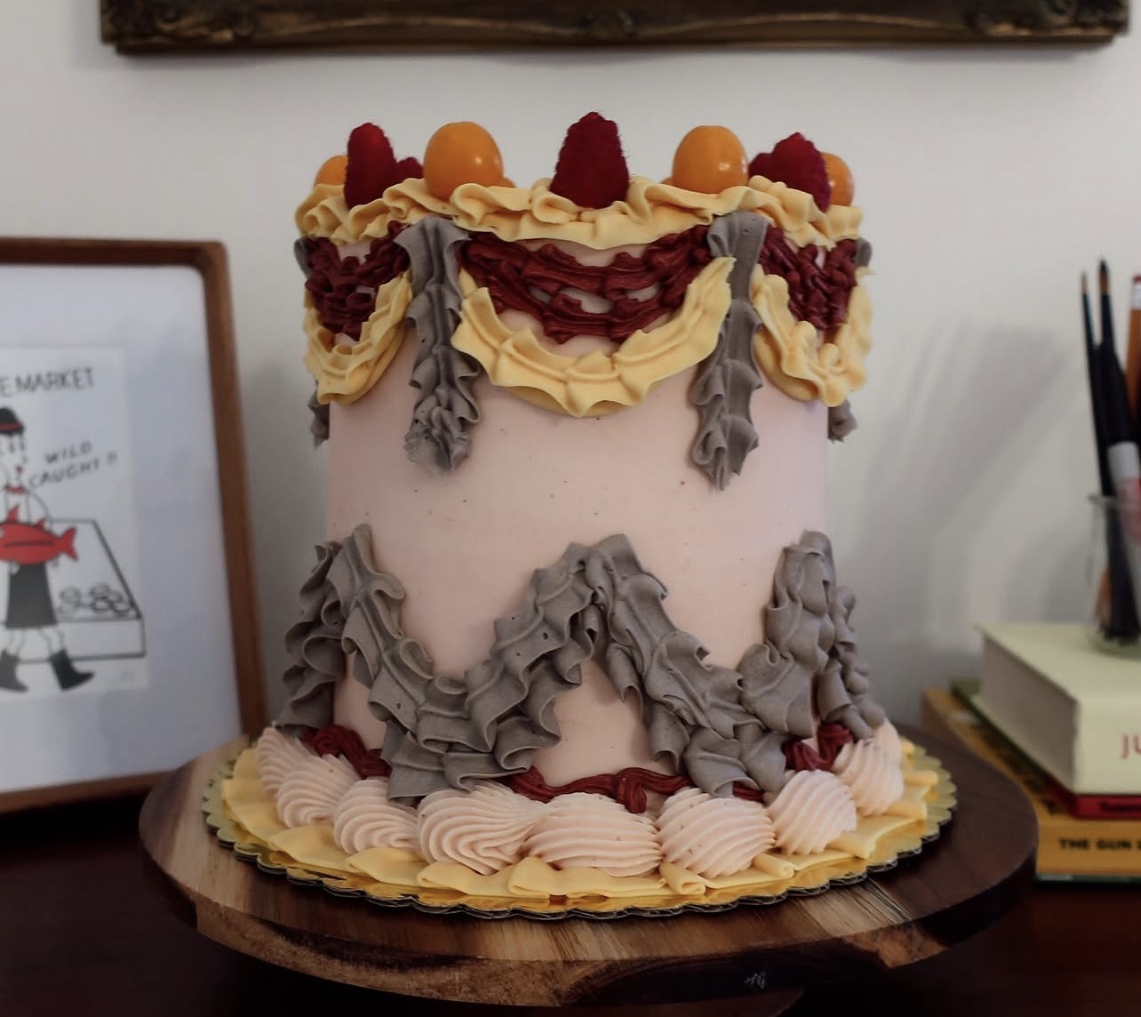Decorated cake with pink frosting, yellow and red fruit, and gray and yellow icing decorations on a wooden surface.