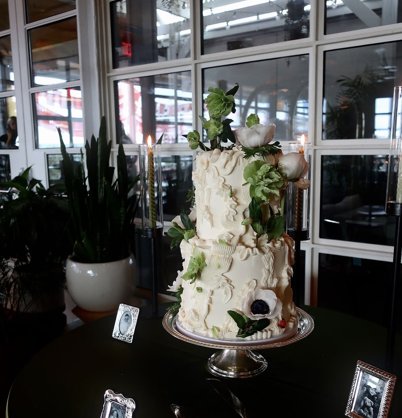 Two-tiered white cake with green leaves and berries on a silver cake stand, in a modern indoor setting.