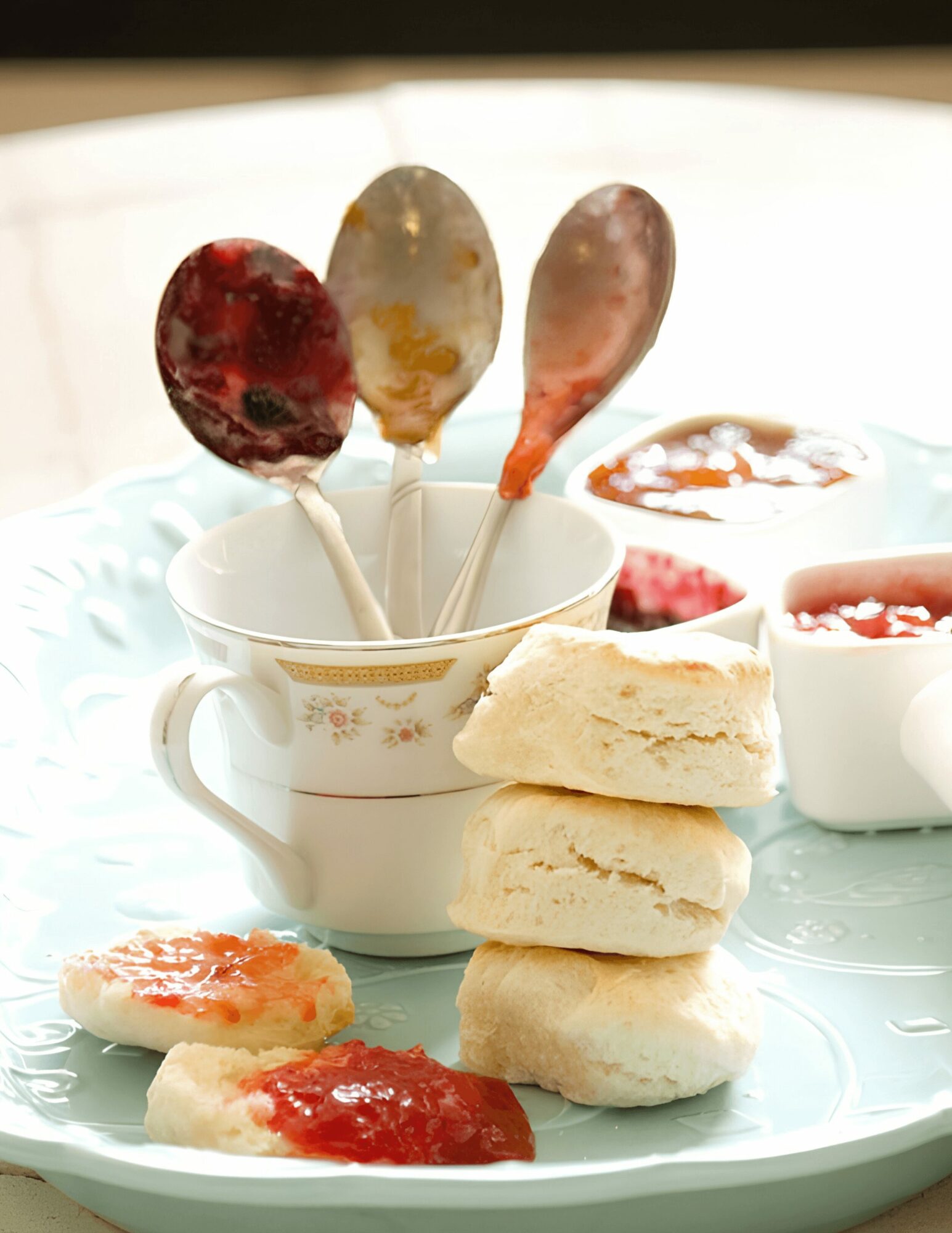 Four scones stacked on a plate with jam, three spoons with jam and jelly in a cup, and small bowls of jam.