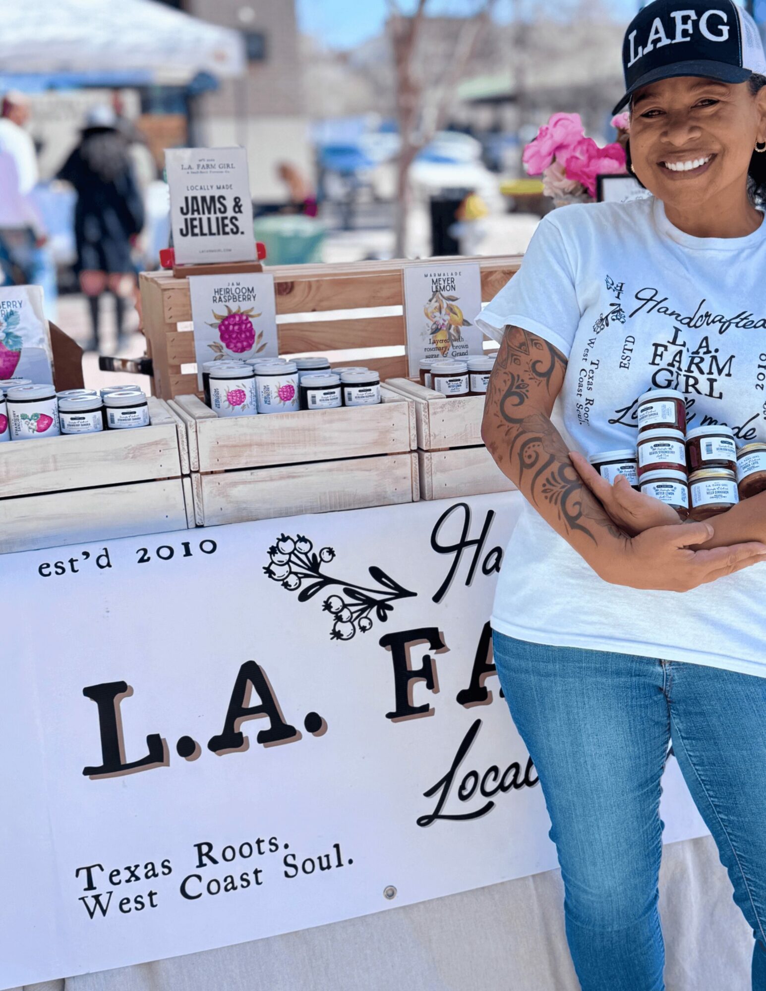 Smiling woman holding jars at outdoor market stall with signs and products, pink flowers in background.