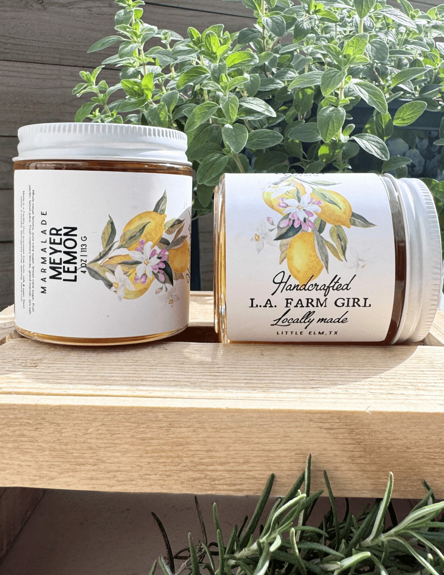 Two jars with lemon and floral labels on a wooden surface, greenery in background.