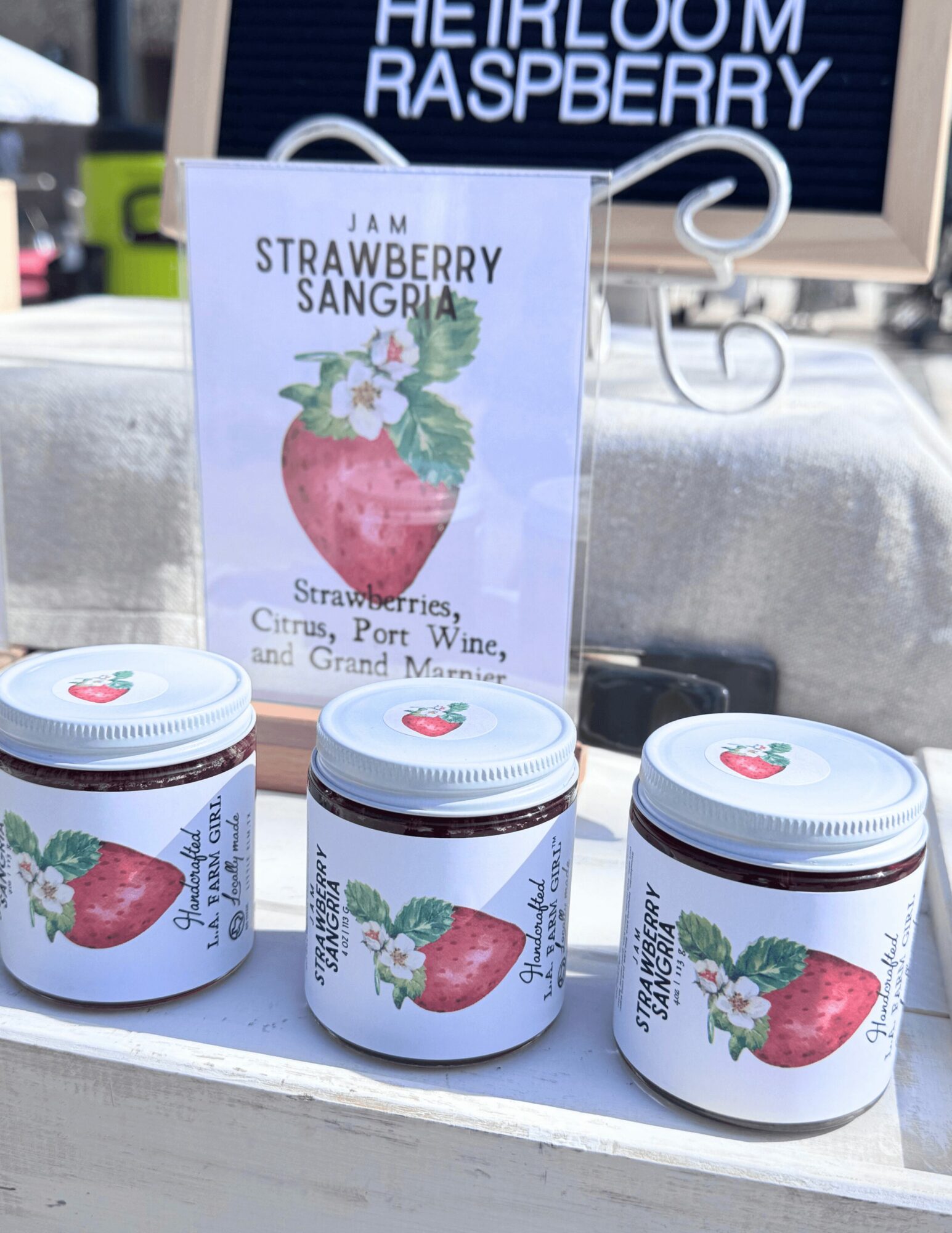 Three jars with strawberry illustrations and labels on a white surface, with a sign in the background reading 'HEIRLOOM RASPBERRY'.