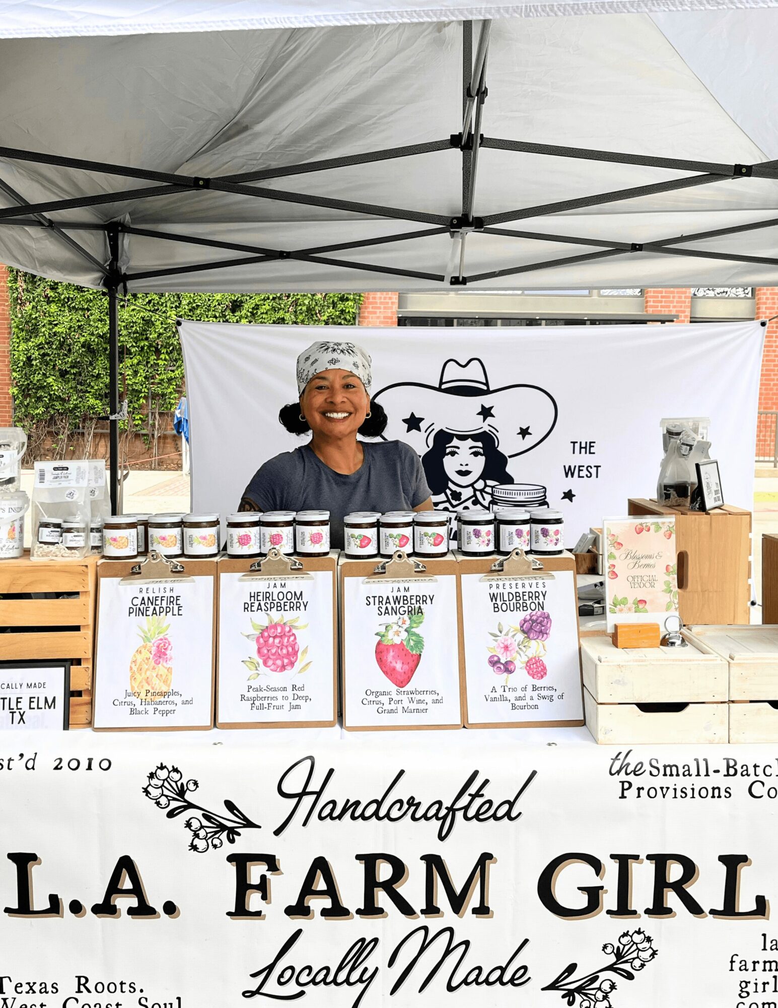 Woman smiling behind a table with jars and signs at an outdoor market stall, canopy overhead, banner in background.