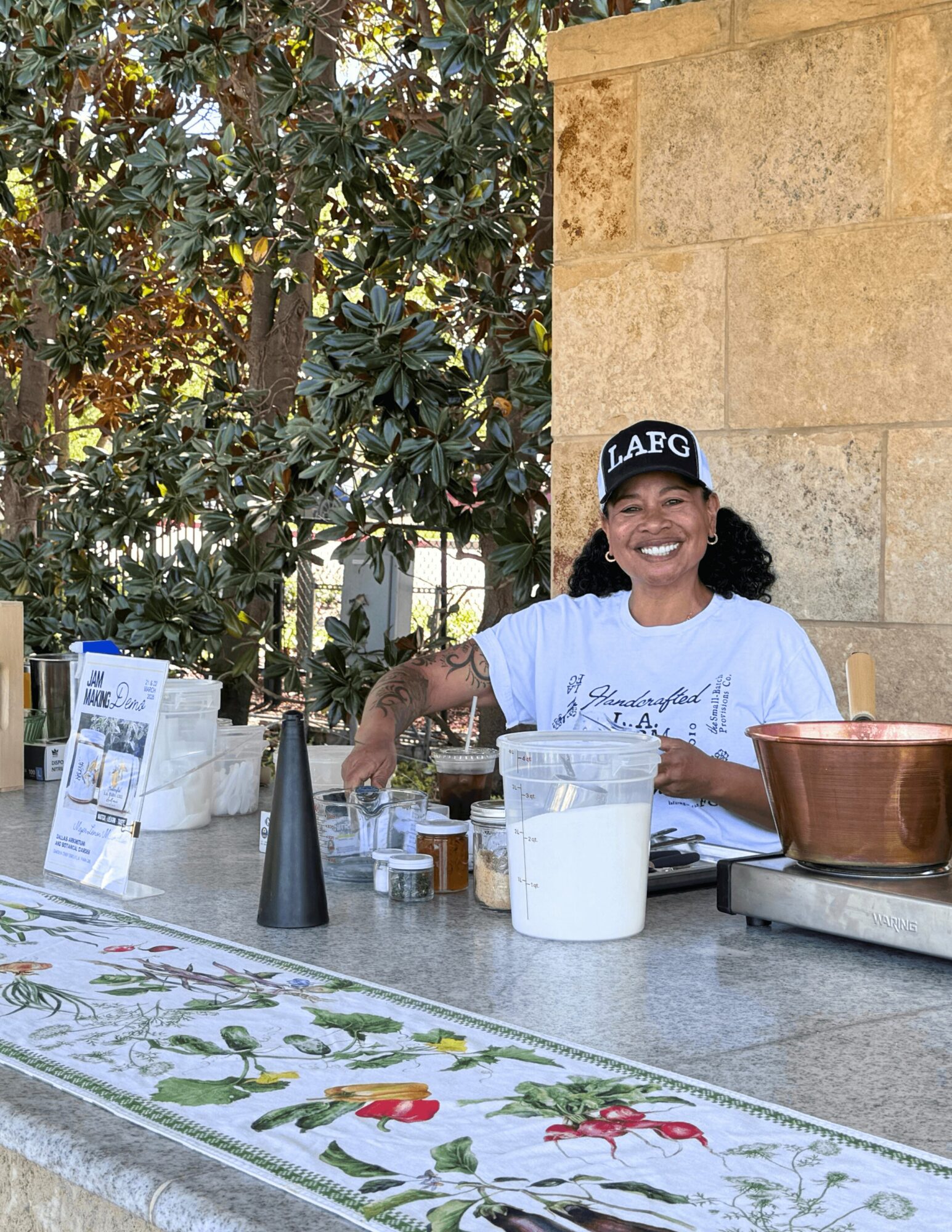 Smiling woman behind a counter with various containers and a copper pot, outdoors with trees and a stone wall.