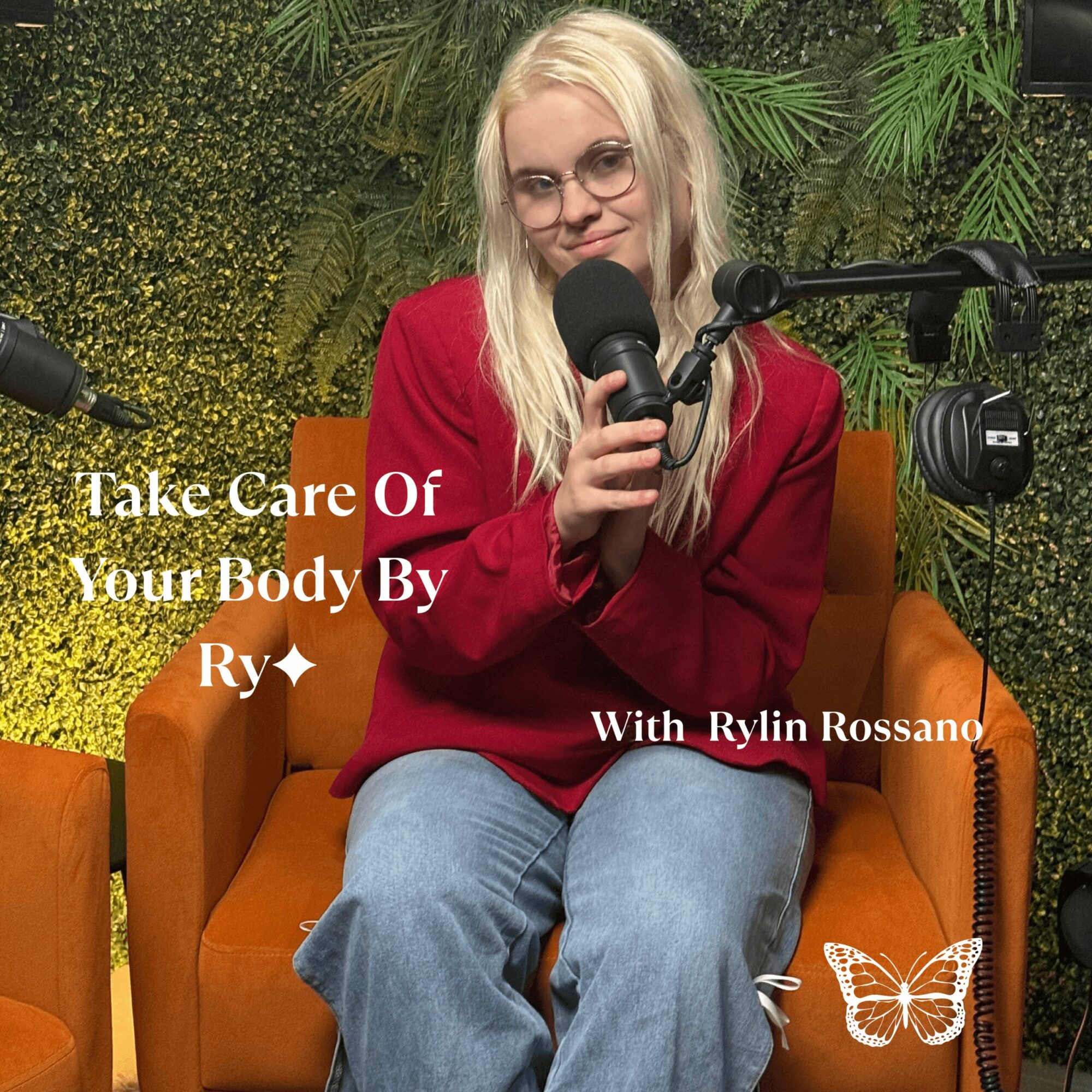 Woman with long blonde hair and glasses sitting on orange chair, holding microphone, green leafy background.