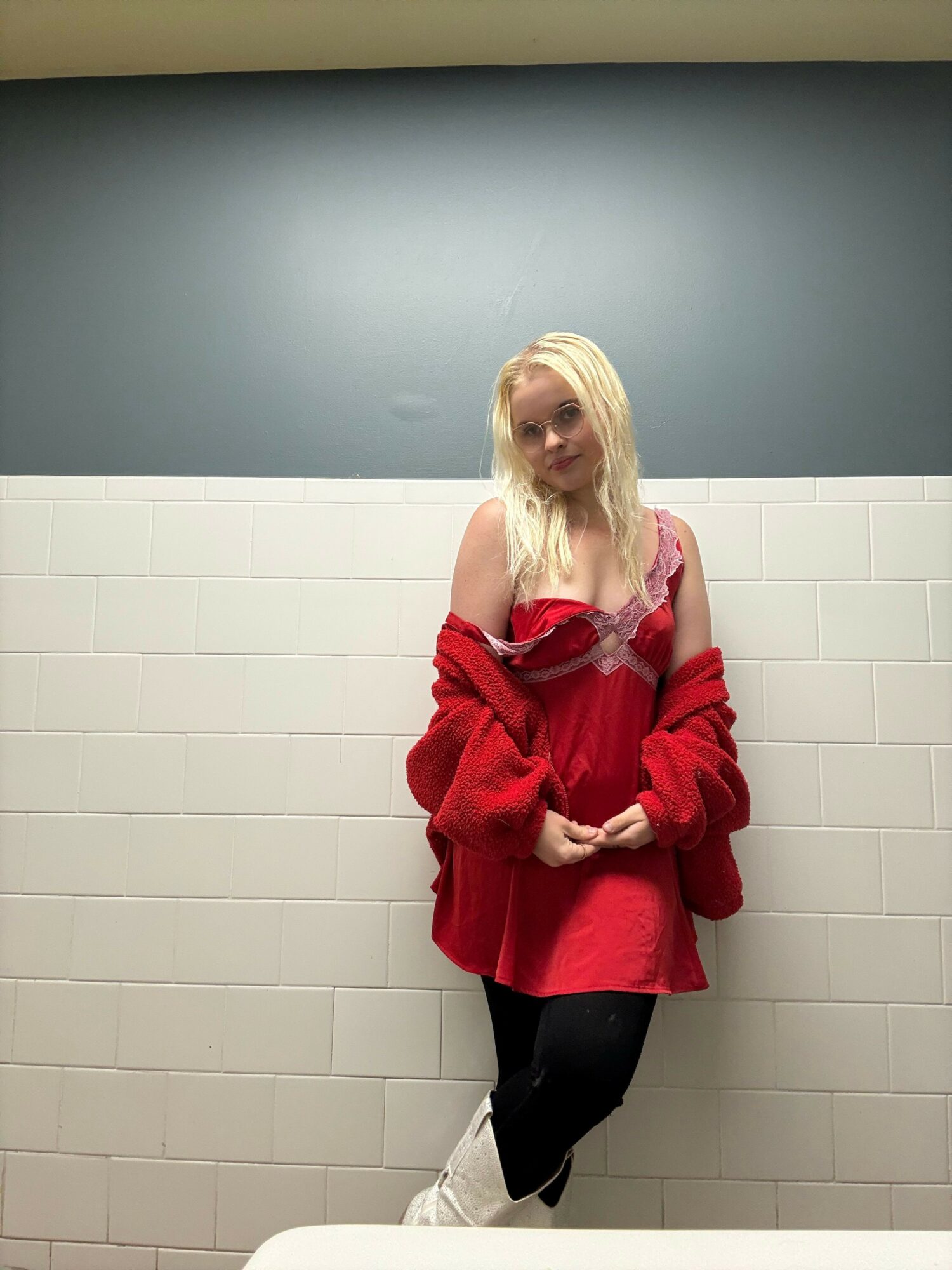 Young woman with blonde hair wearing a red dress and black tights, standing against a wall with white tiles.
