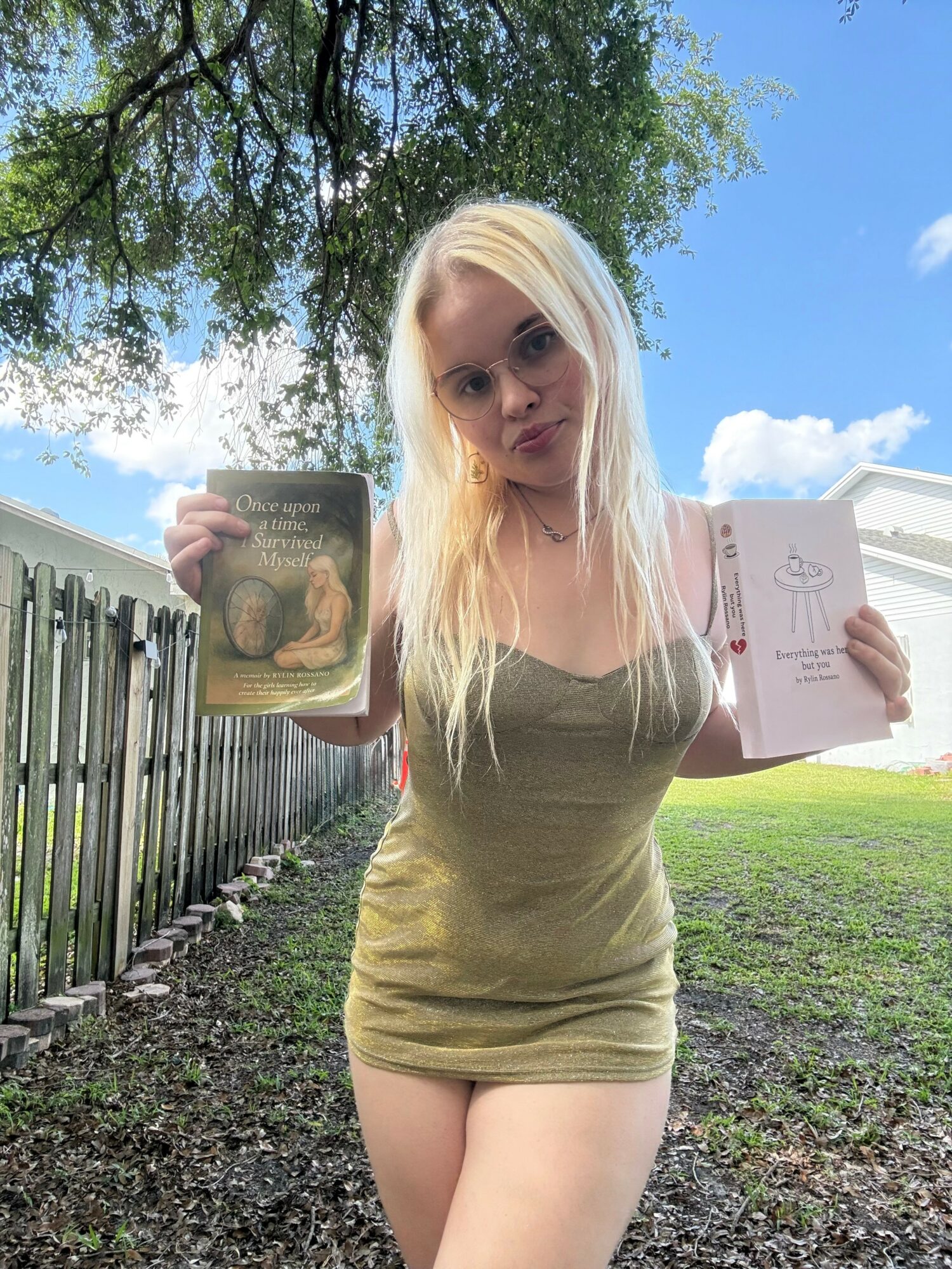 Young woman with long blonde hair holding open book outdoors, standing on grass with fence and trees in background.