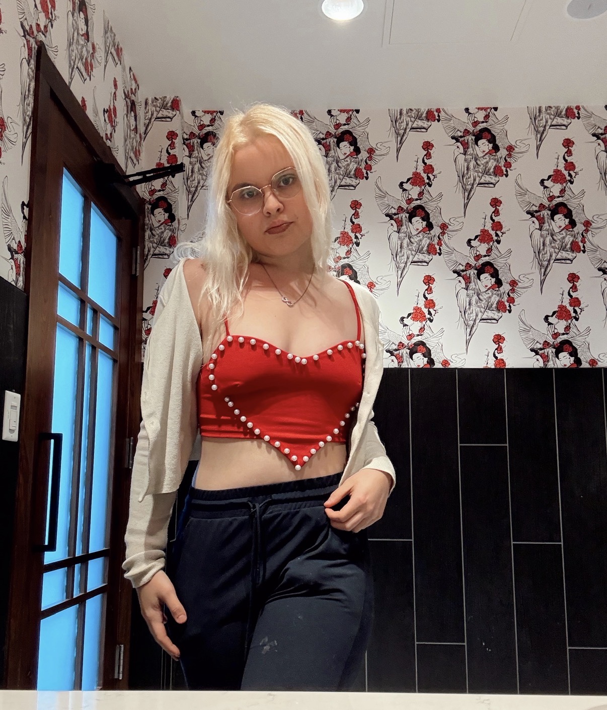 Young woman with blonde hair and glasses standing indoors, wearing a red crop top and dark pants.