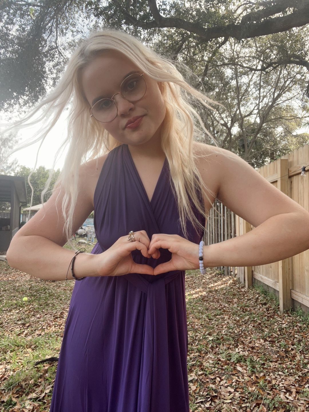 Young woman with long blonde hair wearing sunglasses and a purple dress outdoors, making a heart shape with her hands.
