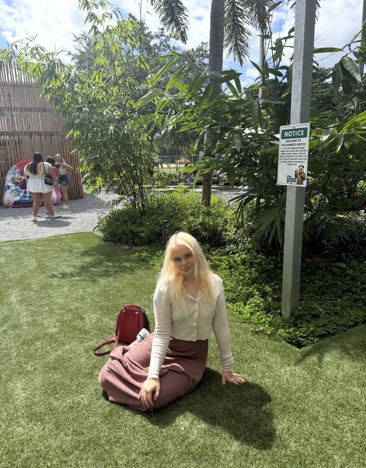 Woman with blonde hair sitting on grass in a garden area, surrounded by trees and plants, with people in the background.