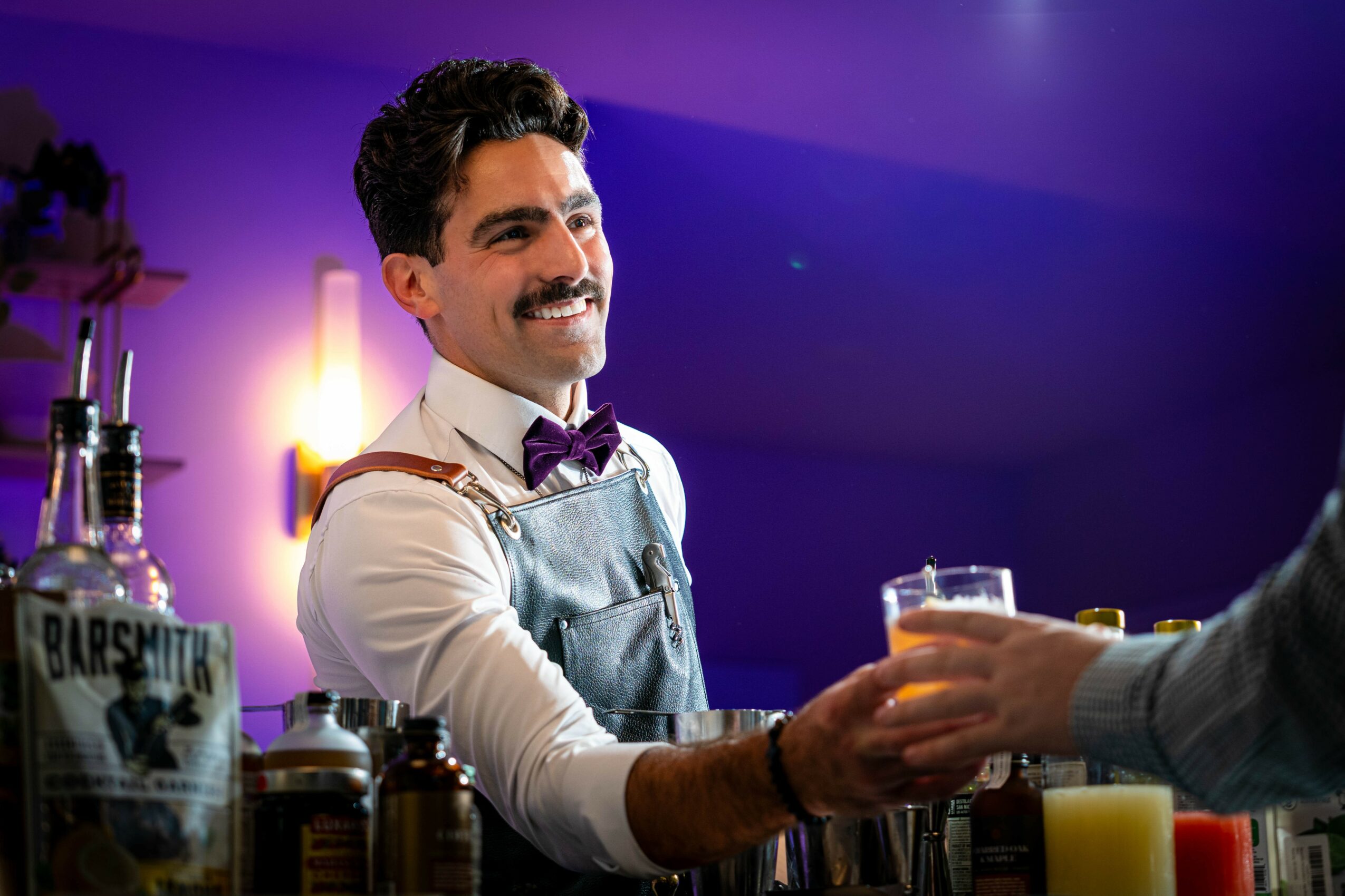Bartender smiling and handing a drink to a customer in a bar setting.