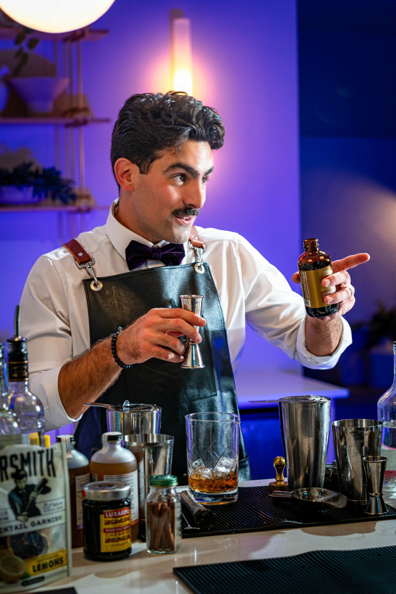 Bartender with dark hair, mustache, wearing apron and bow tie, holding a bottle and glass, behind bar with bottles and glasses.