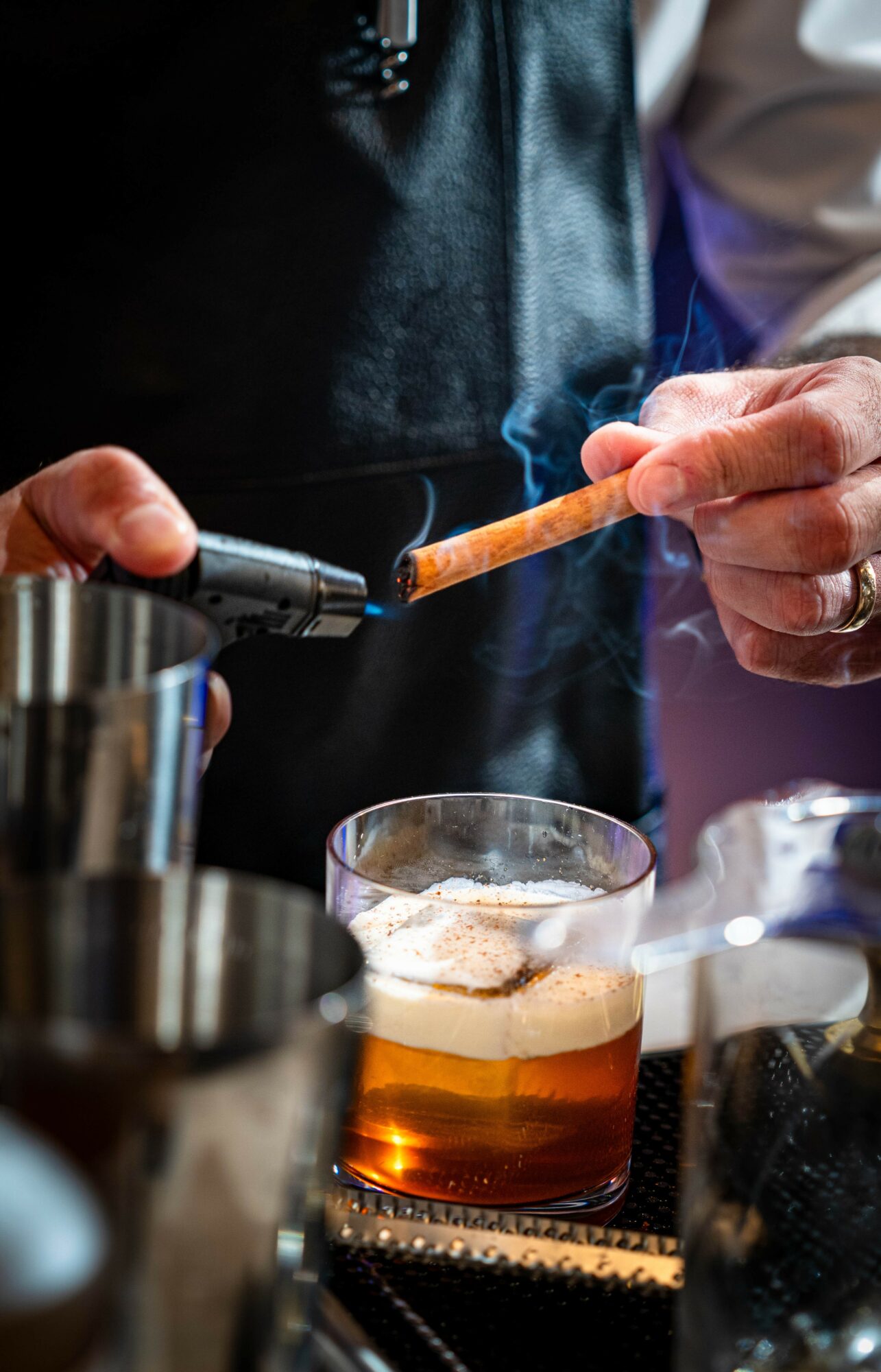 Person holding a lit cigarette over a glass of beer with foam, surrounded by bar tools.